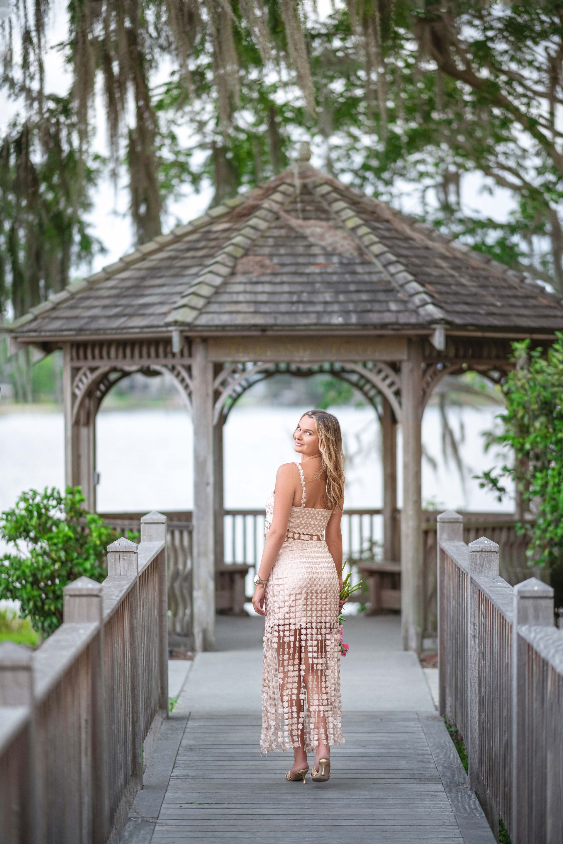 Woman in a sparkly dress looks back over her shoulder while standing on a wooden bridge, gazebo in the background.