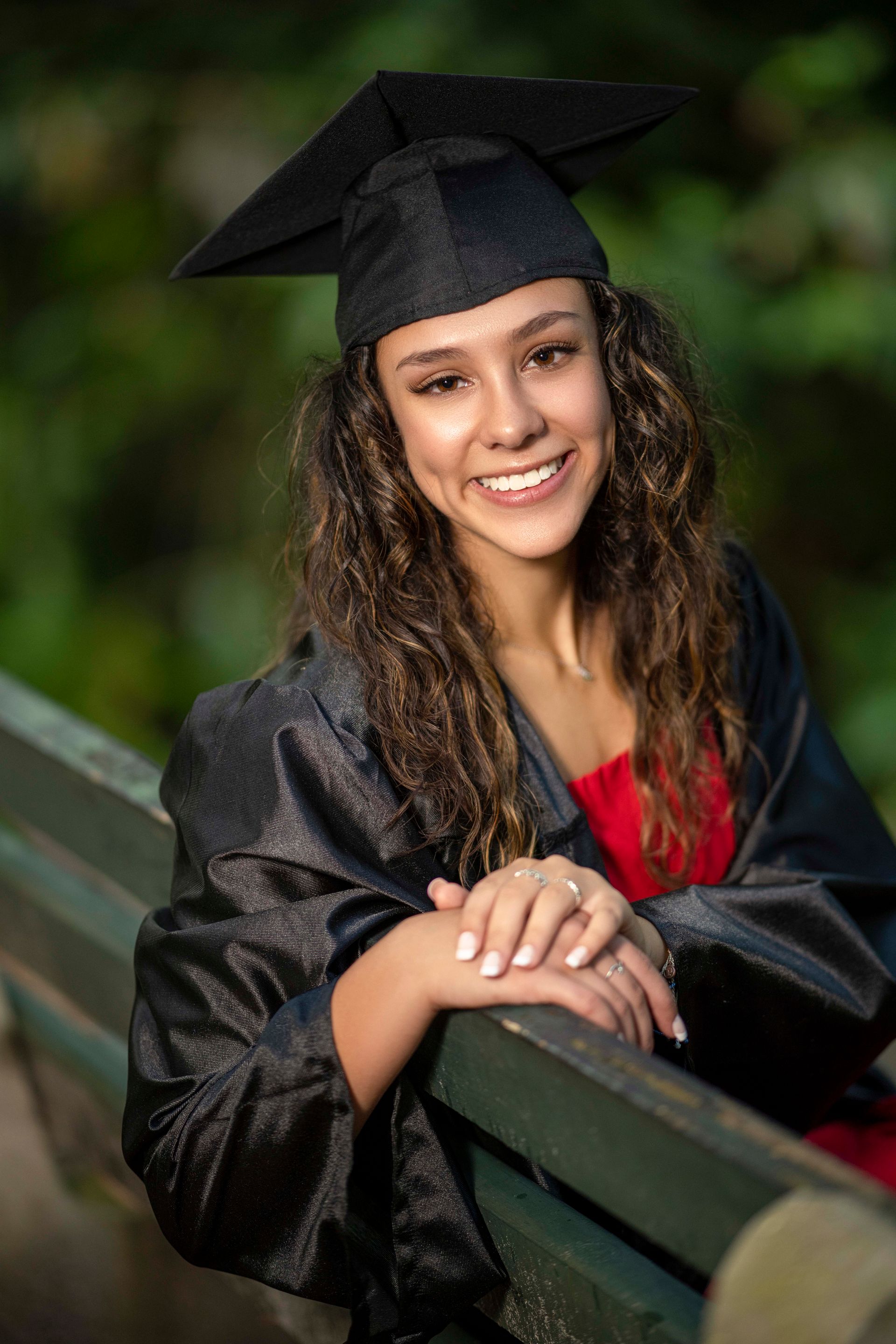 Woman in graduation cap and gown smiles, leaning on a bench in an outdoor setting.