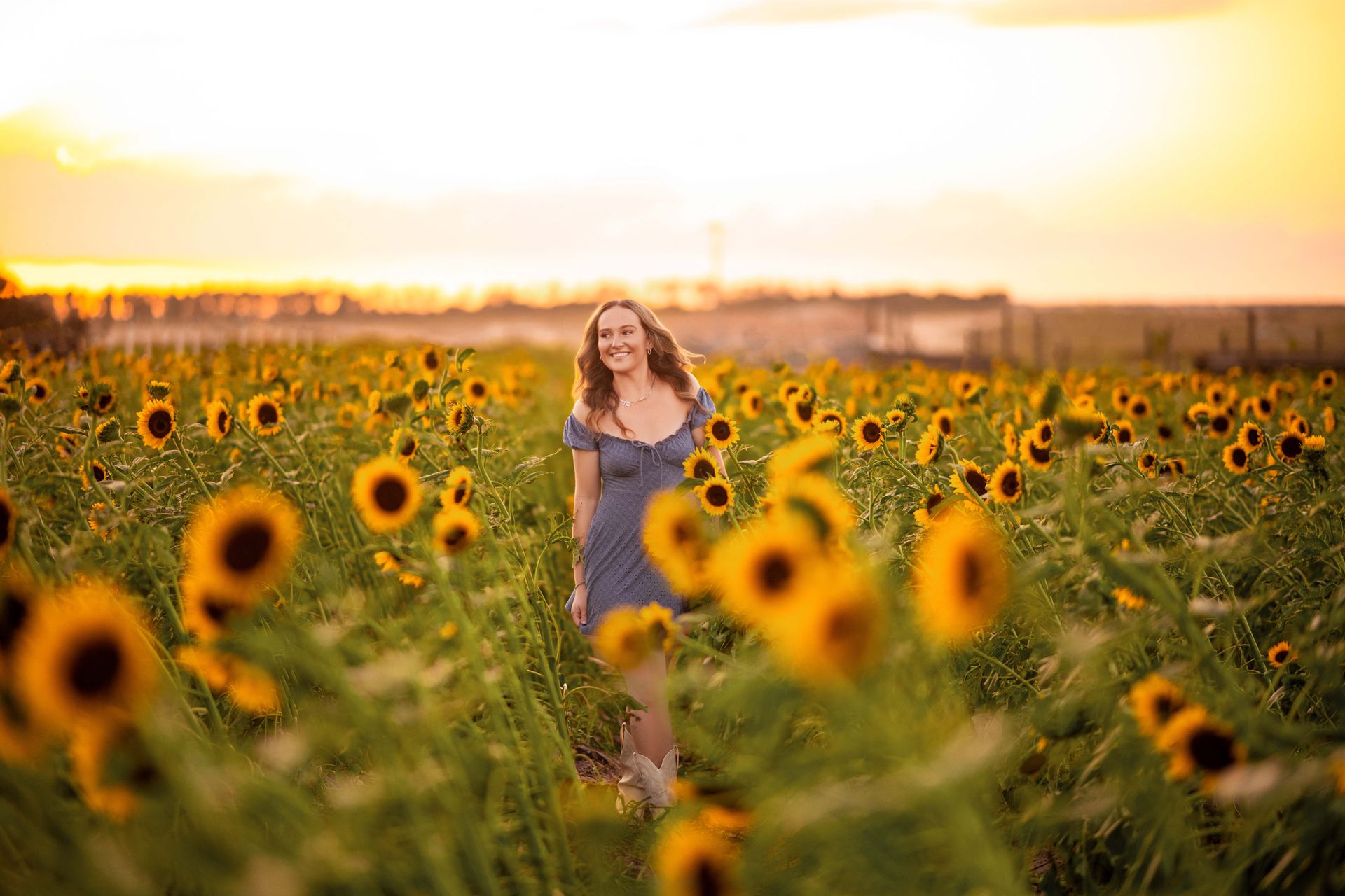 Woman smiling in a field of sunflowers at sunset, wearing a blue dress.