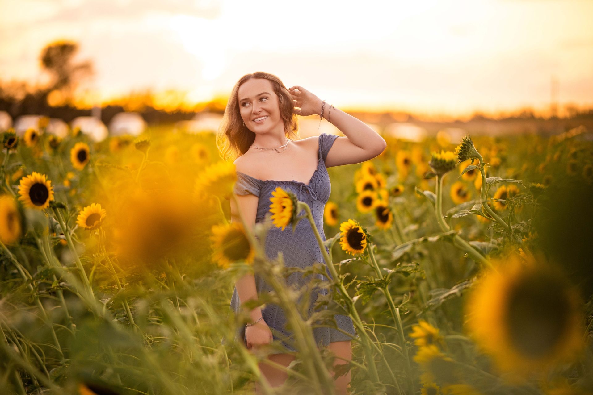 Woman in blue dress smiles in a sunflower field at sunset, touching her hair.