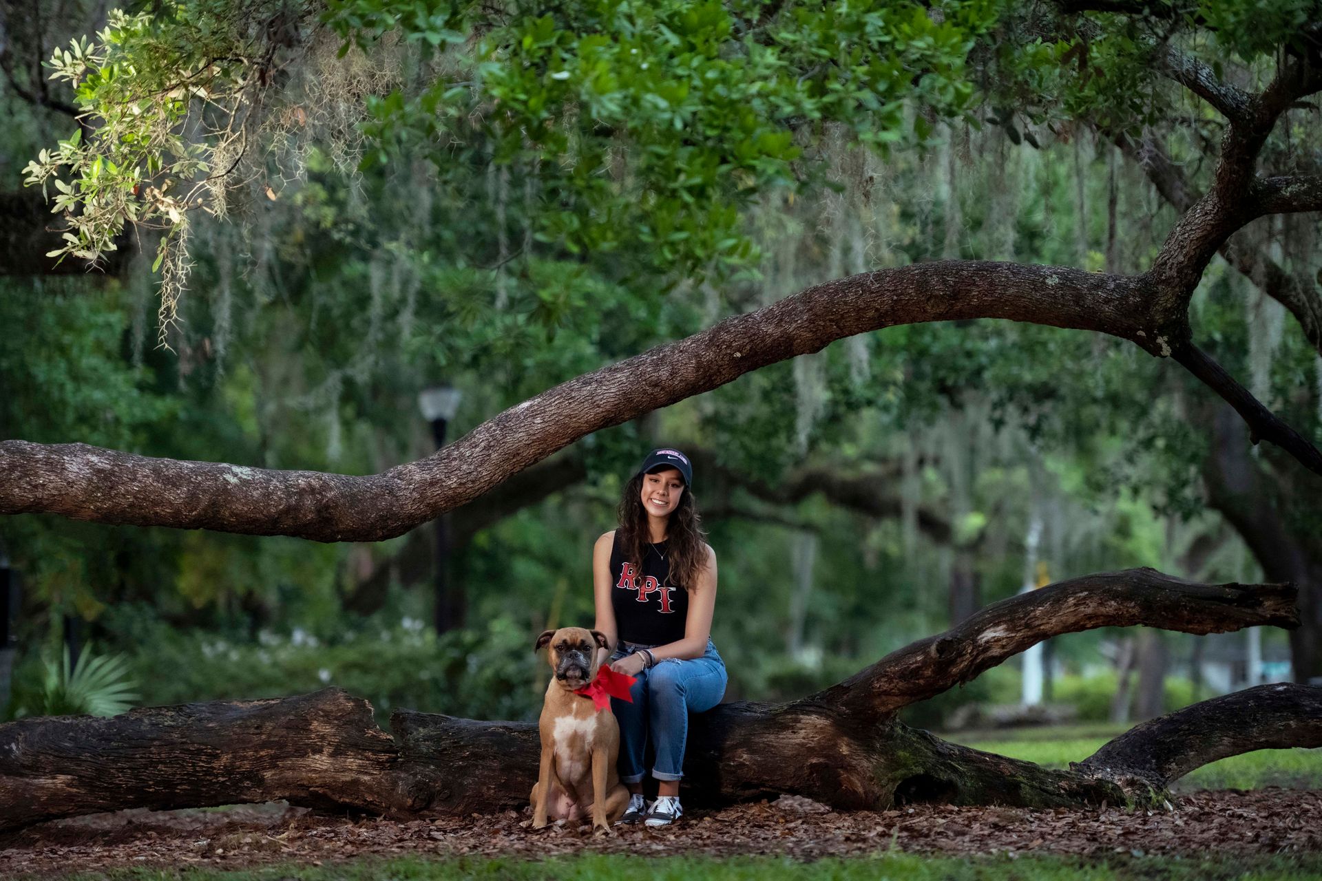 Woman and dog sit under large tree, wearing a black hat and shirt, and blue jeans.