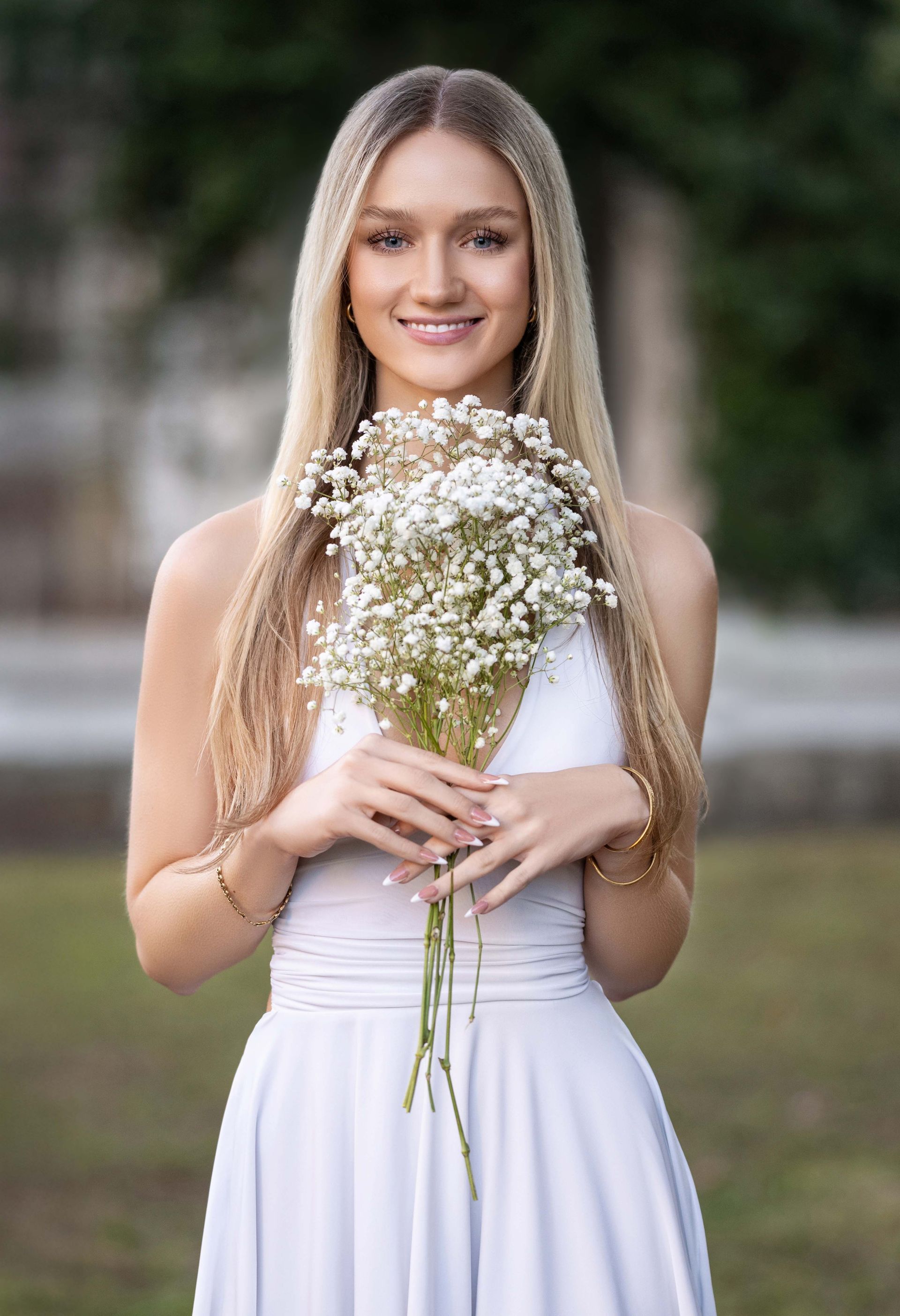Woman with long blonde hair, holding a bouquet of white flowers, smiling, wearing a white dress, outdoors.