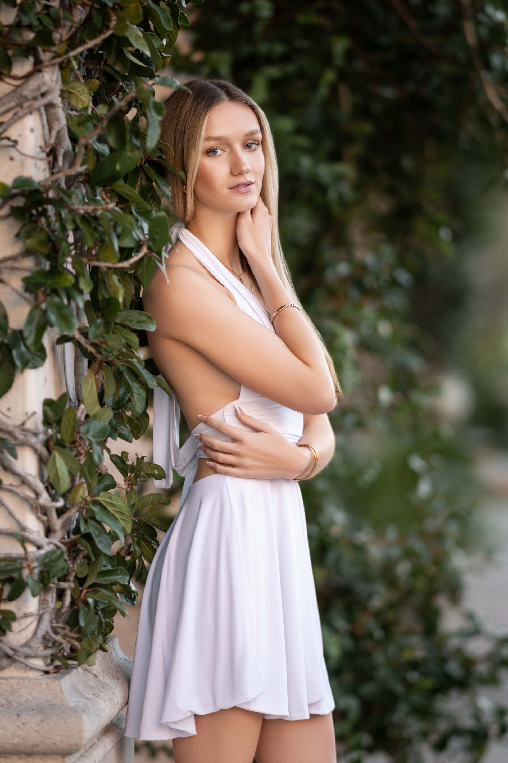 Blonde woman in a white dress poses next to a column covered in ivy; outdoor setting.