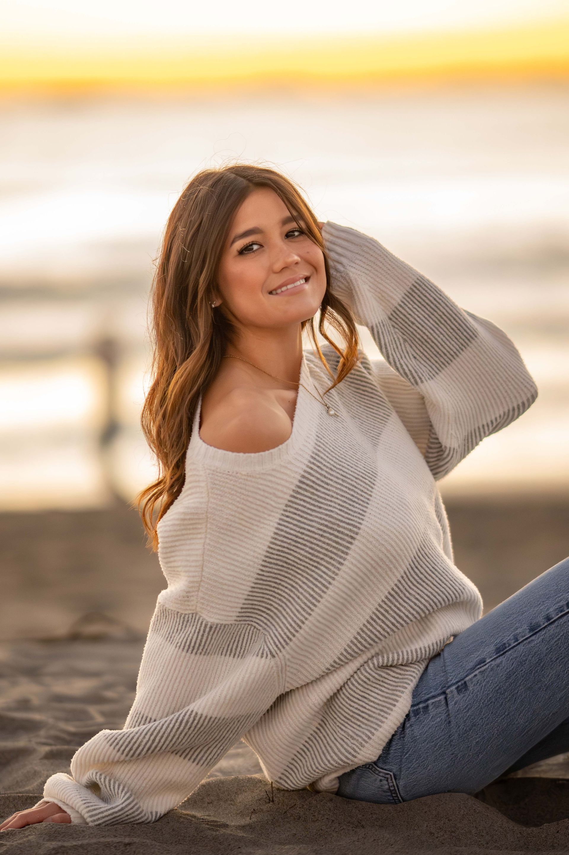 Young woman with long hair smiles, wearing a sweater and jeans, sitting on the beach at sunset.