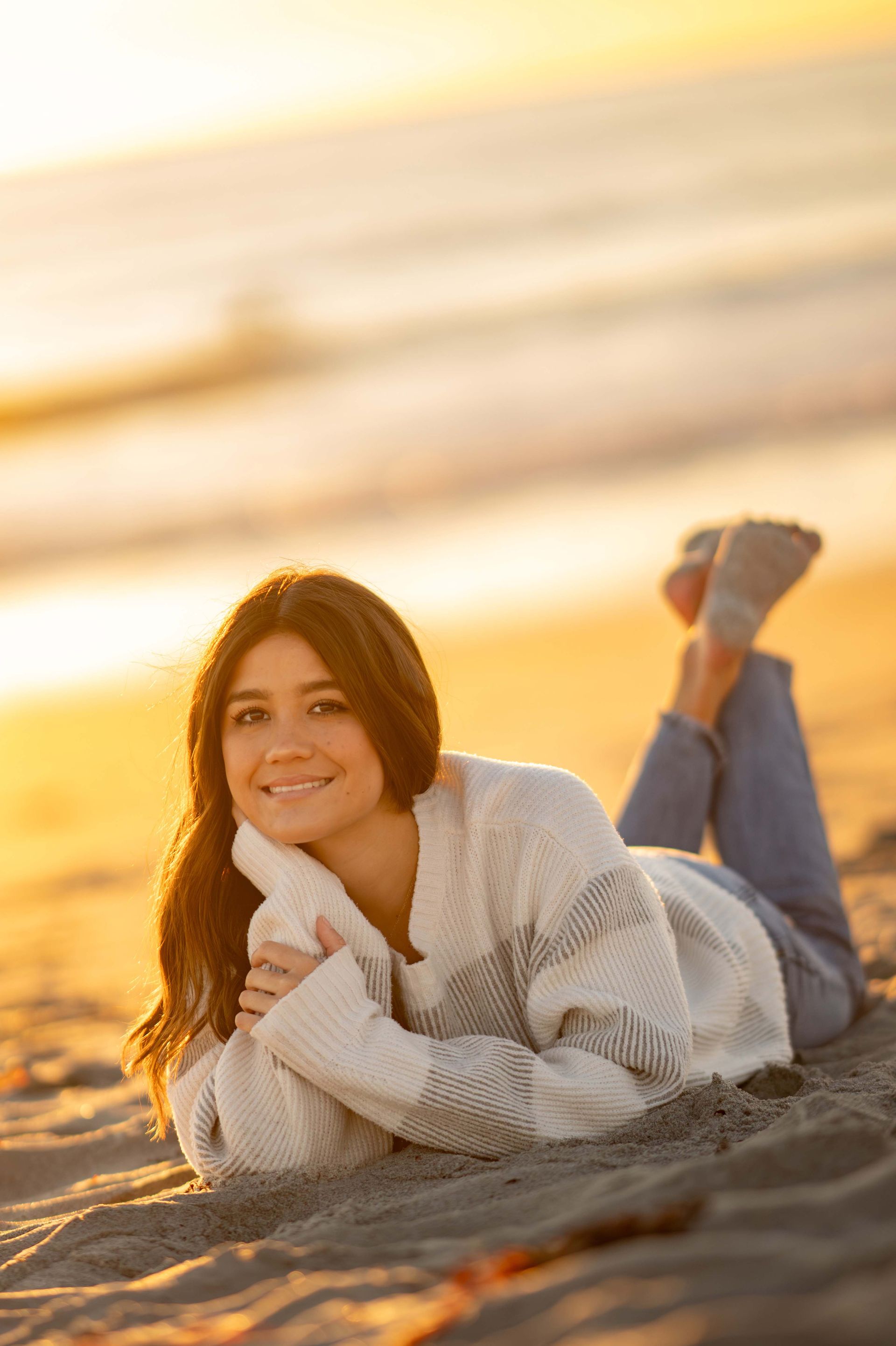 Woman smiles while lying on beach, wearing sweater and jeans, during sunset.