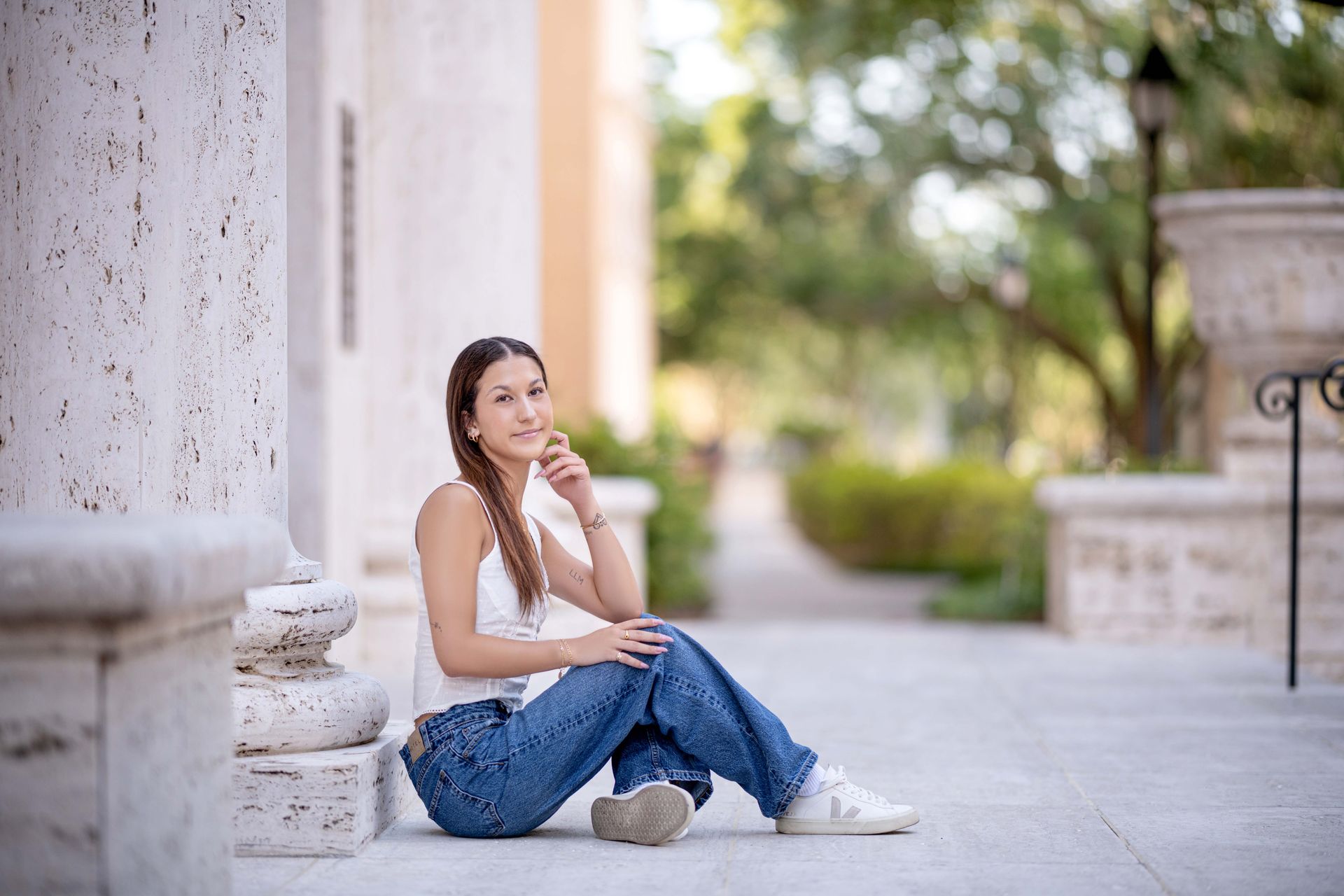 Woman in jeans and white top sitting by a pillar outside, smiling.