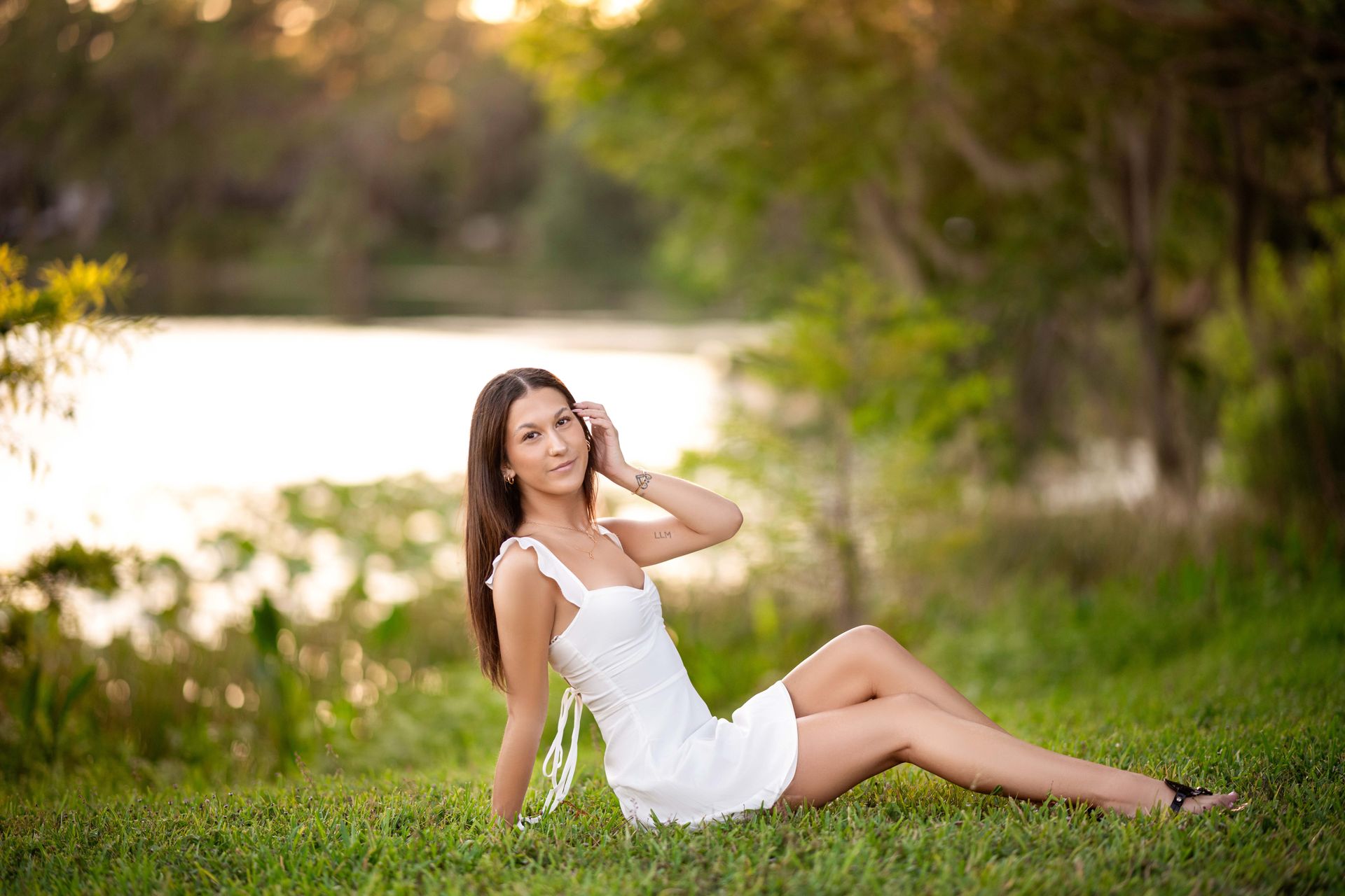 Woman in a white dress sitting on grass by a lake, smiling. Sunlight and trees in the background.