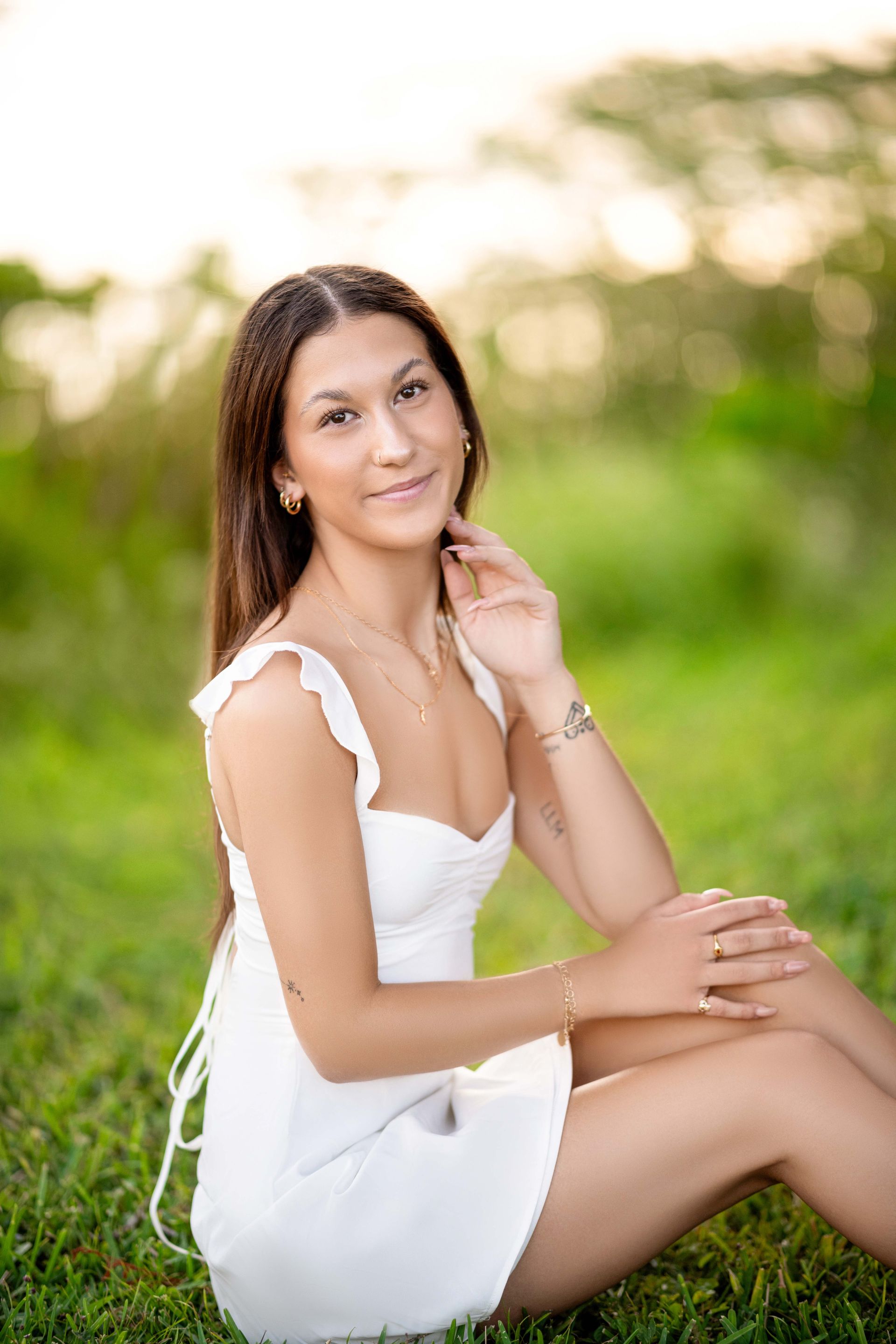 Woman with long dark hair, wearing a white dress, sitting in a grassy field, smiling.