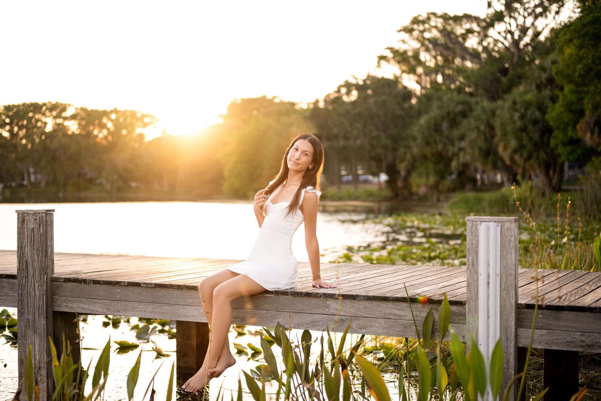 Woman in white dress sitting on a wooden dock by a lake at sunset, smiling at the camera.