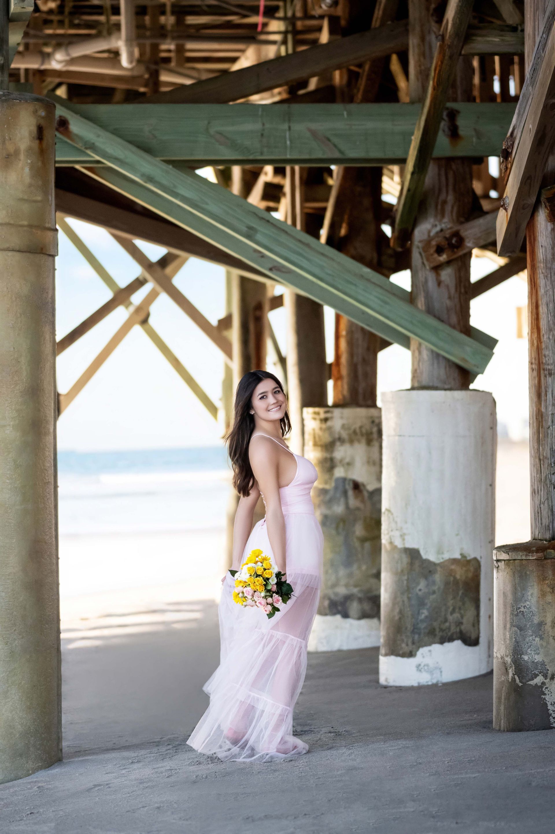 Woman in pink dress smiles, holds flowers, stands under pier; beach in background.
