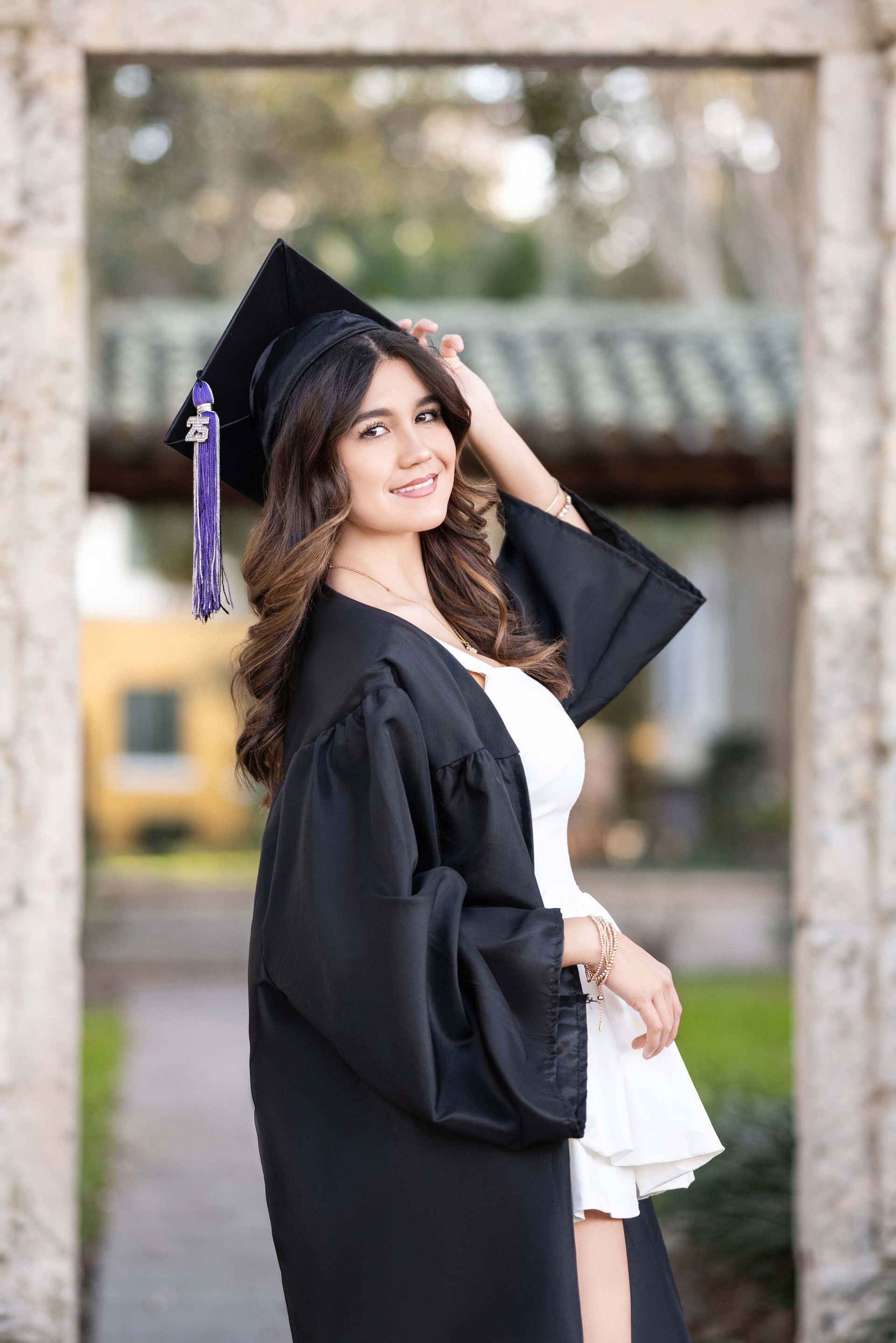 Woman in cap and gown smiles, posing in a stone archway, white dress.