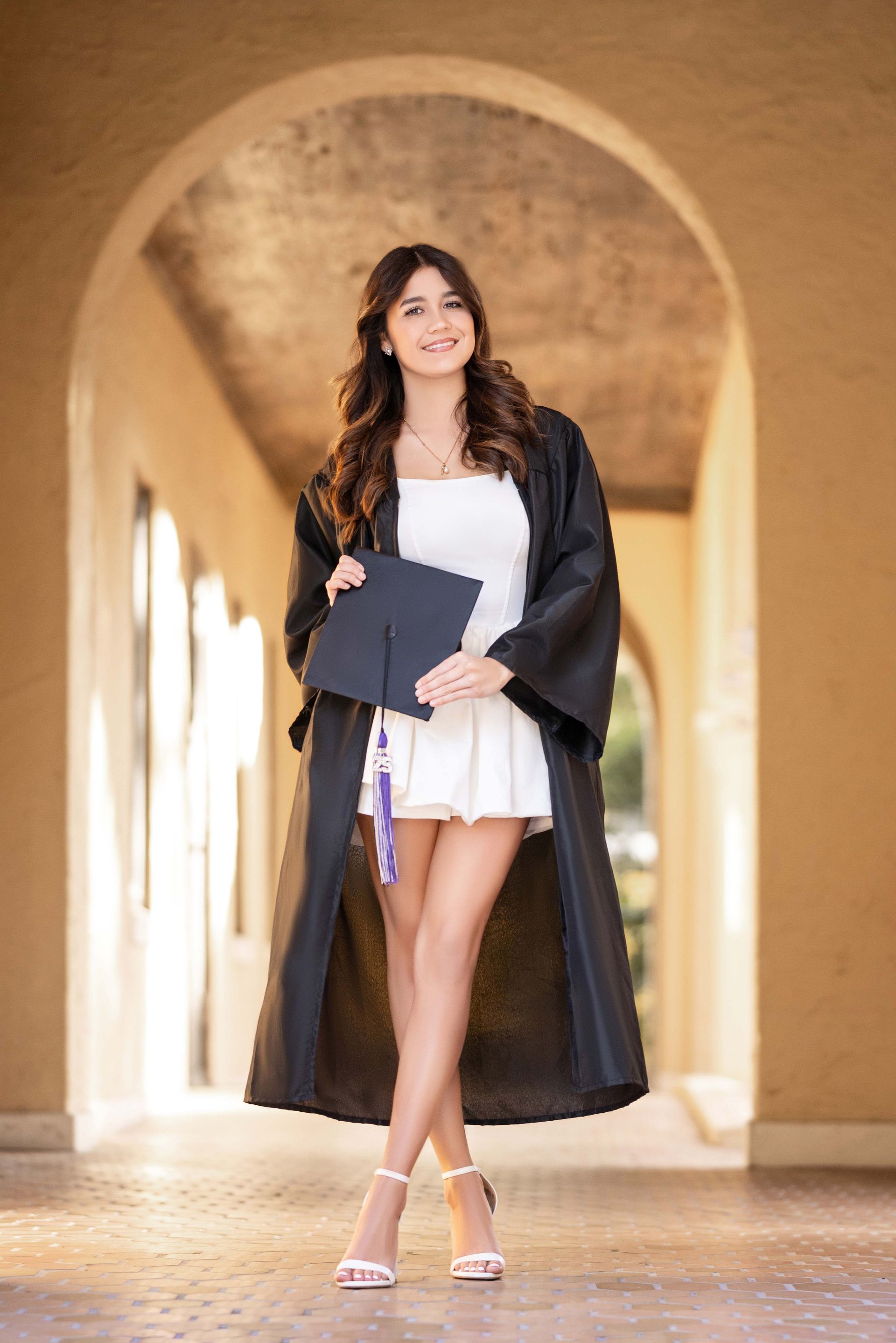 Young woman in graduation robe holding cap, smiling, standing in a hallway.
