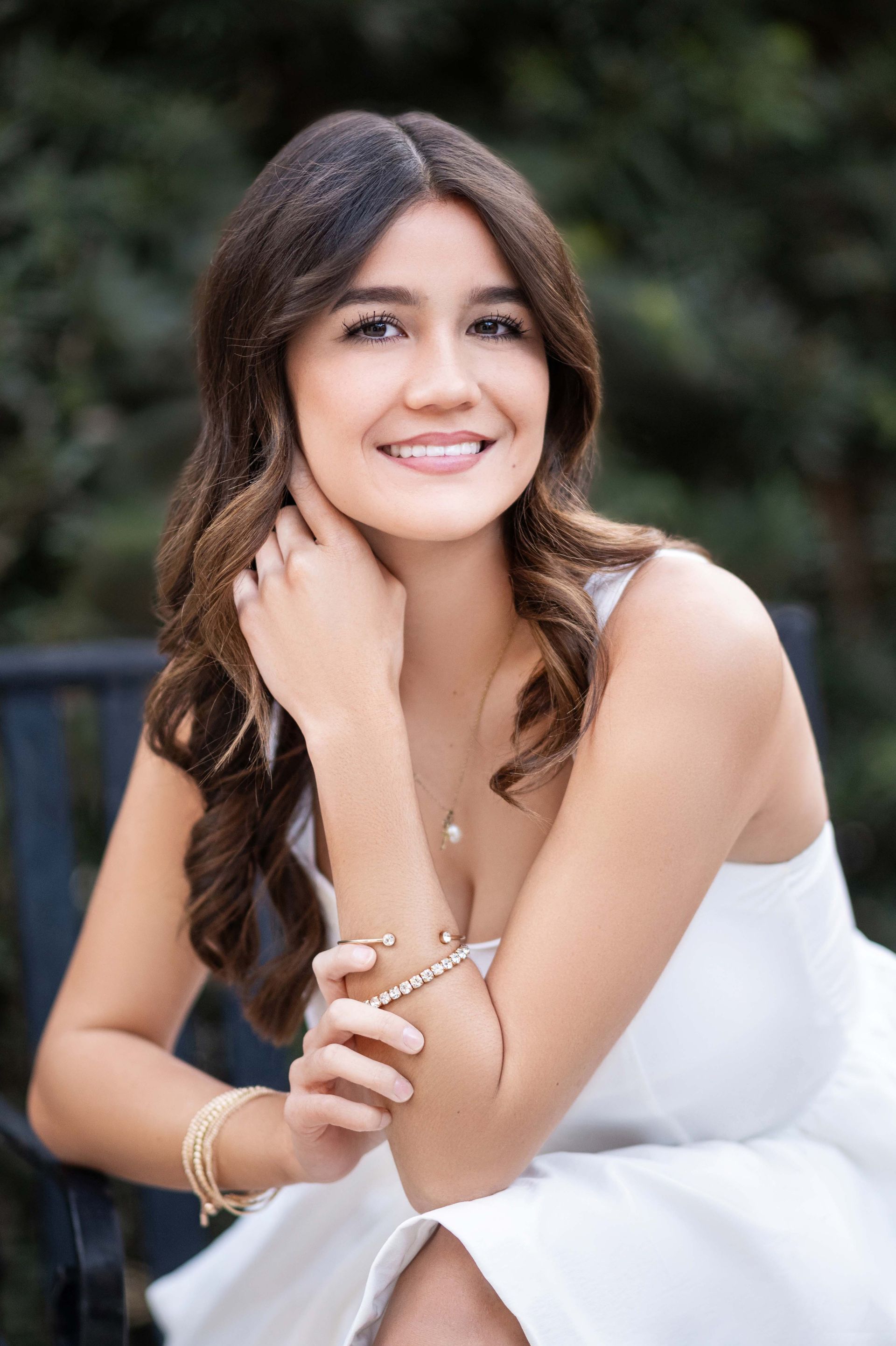 Woman with dark hair, in white dress, smiling outdoors, wearing jewelry.