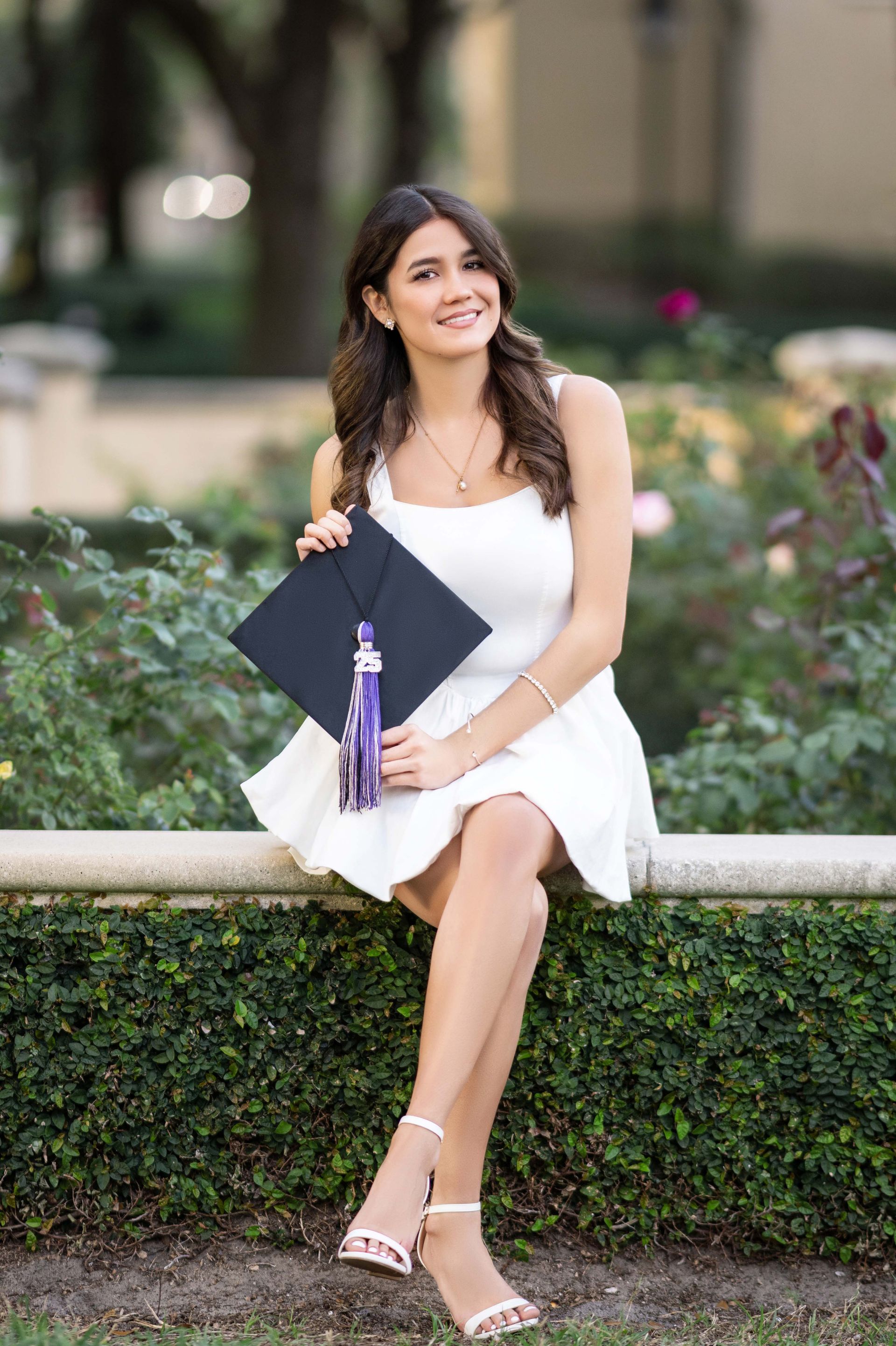 Young woman in white dress, graduation cap, sitting on a wall, smiling in a garden.