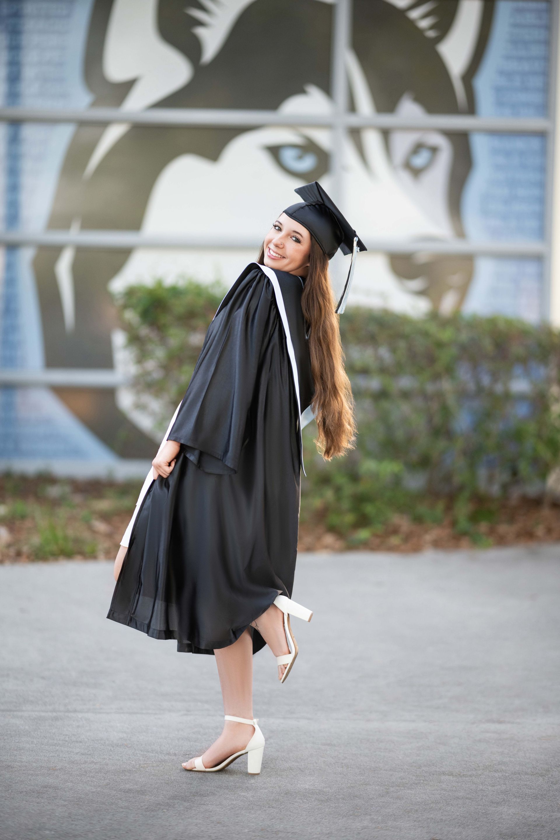 Woman in graduation gown poses in front of a building with a wolf logo.