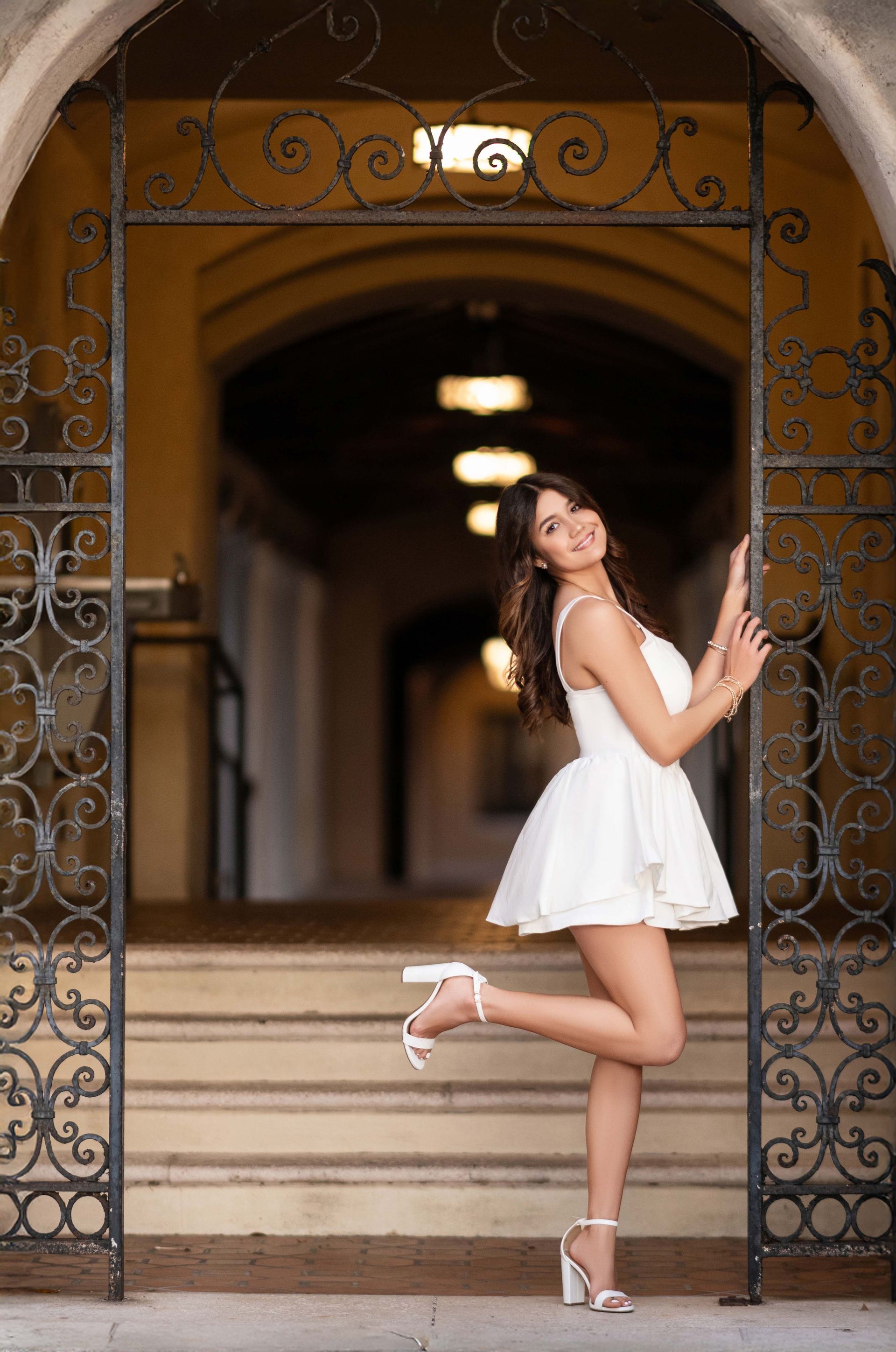 Woman in white dress poses by an ornate gate in a sunlit building.