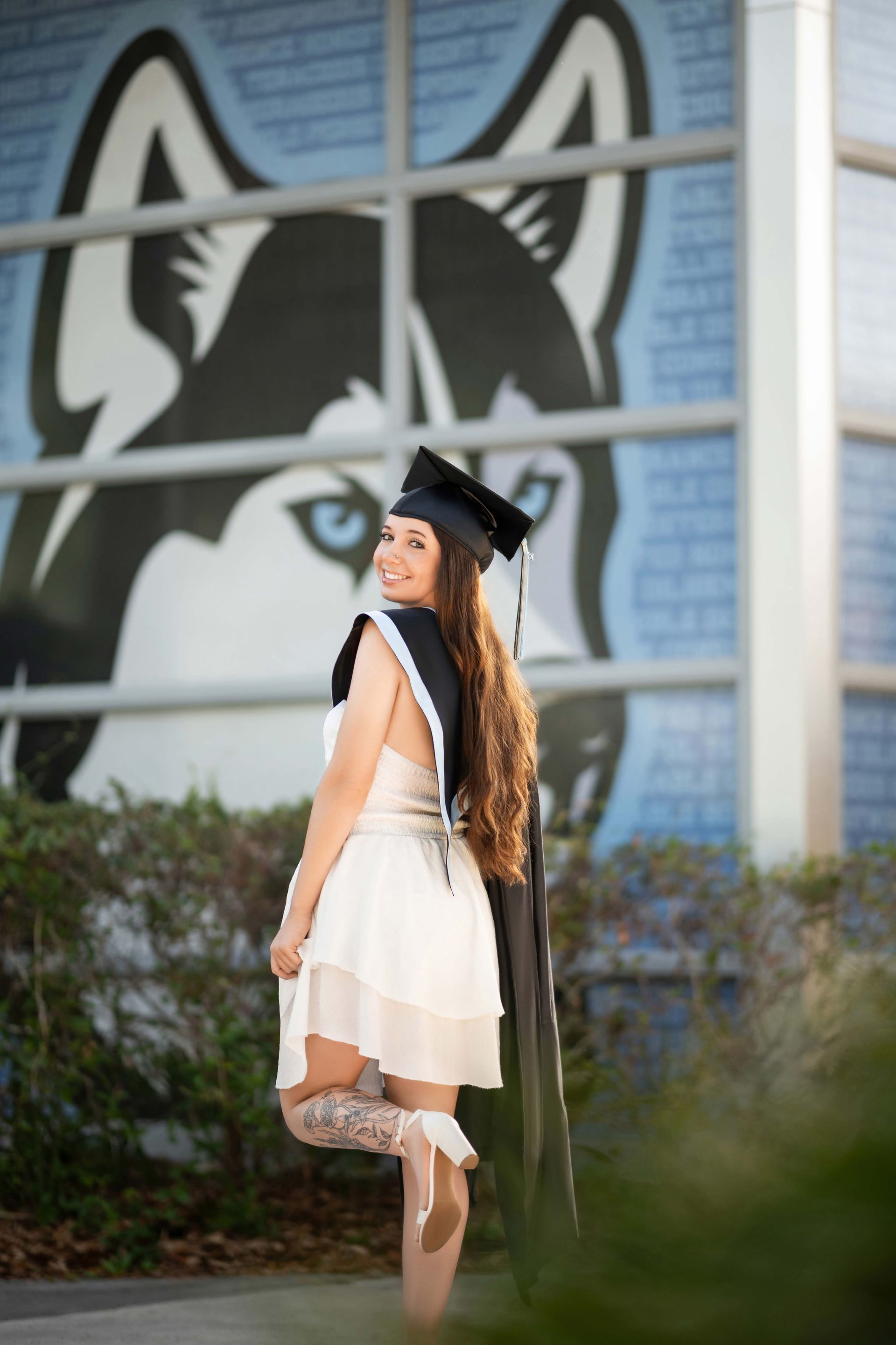 Woman in graduation cap and gown smiles, posing in front of a husky logo on a building.