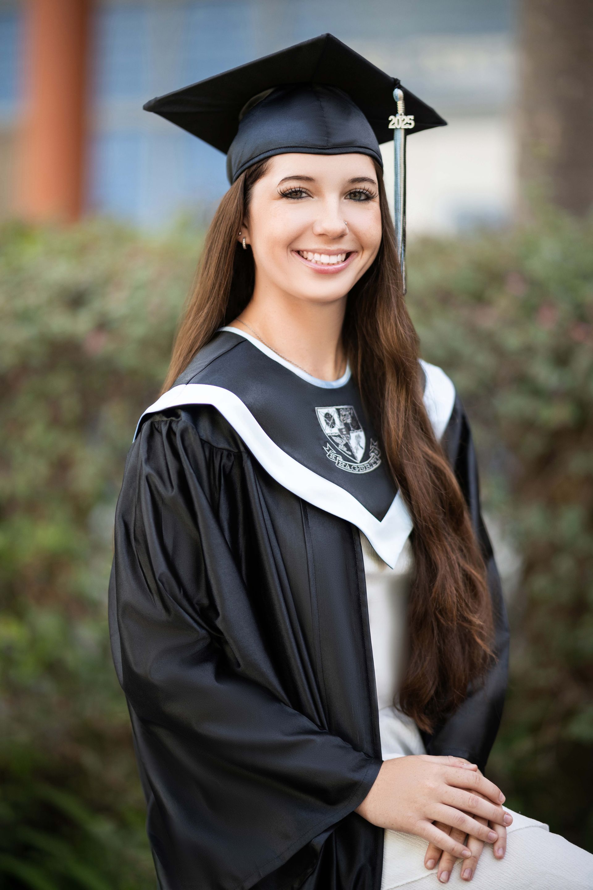 Young woman in graduation attire, smiling, in front of greenery.