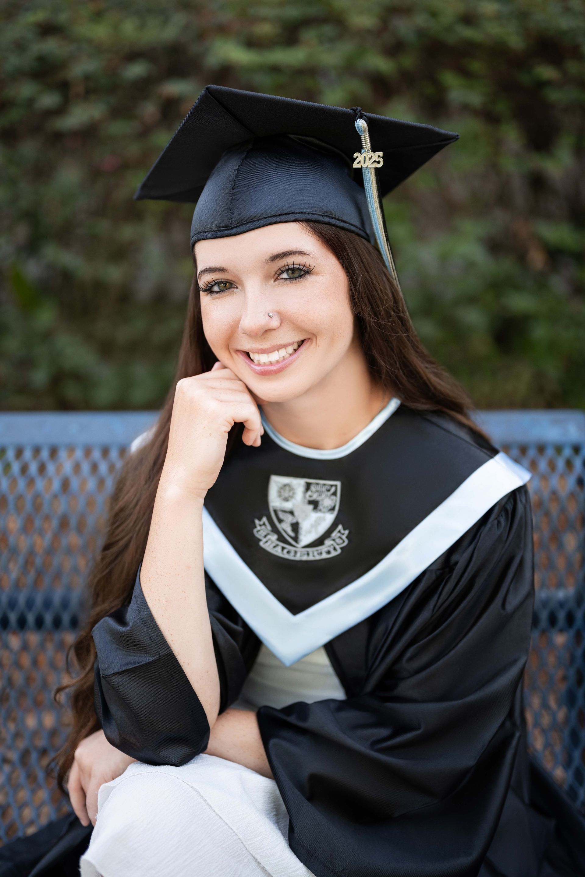 Woman in graduation gown smiling, sitting outside, hand on chin, long brown hair, black cap.