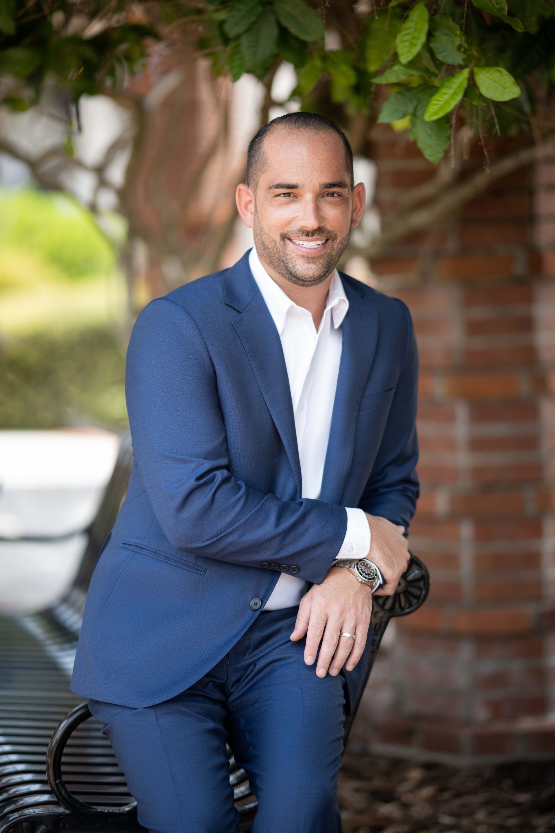 Man in blue suit smiles while leaning on a bench; outdoor setting with brick wall and greenery.