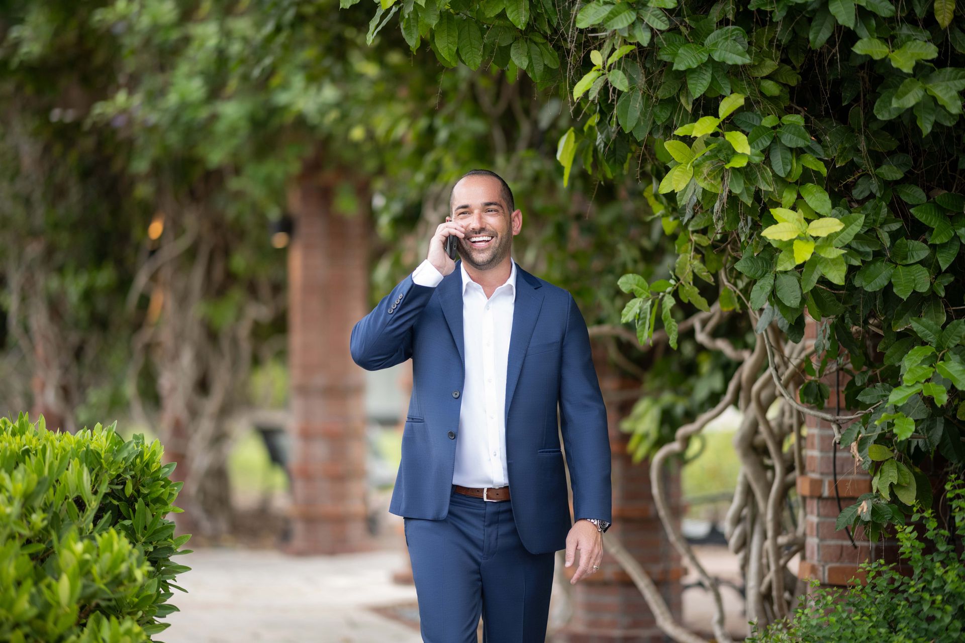 Man in blue suit walking outdoors, talking on phone, smiling near greenery.