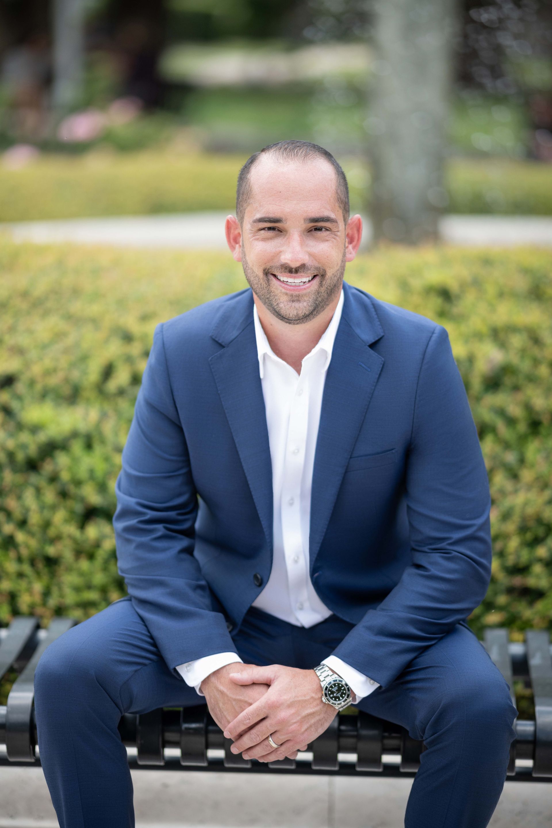 Man in blue suit sits on bench, smiling at the camera outdoors.