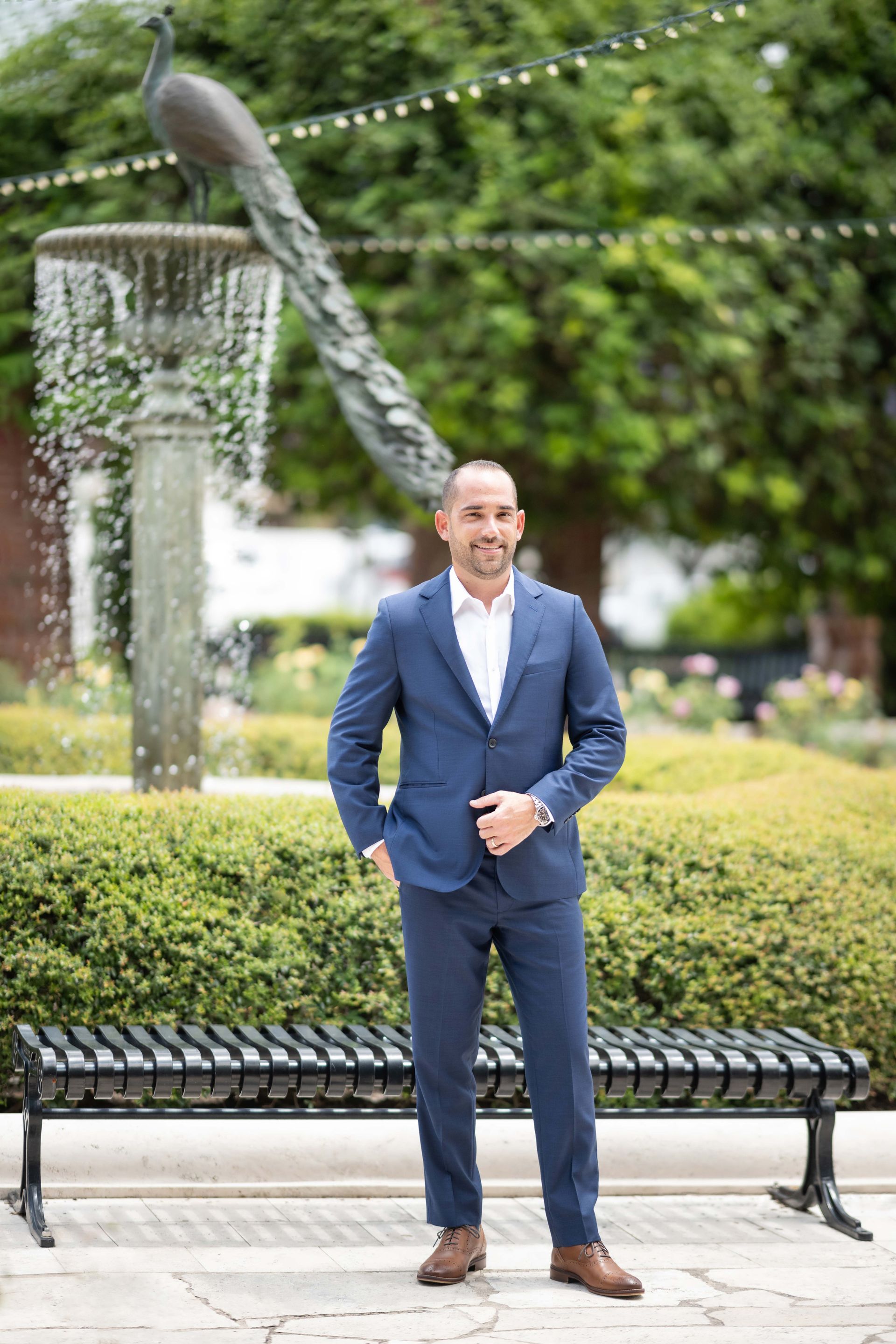 Man in blue suit stands by a fountain with a peacock statue and a bench.