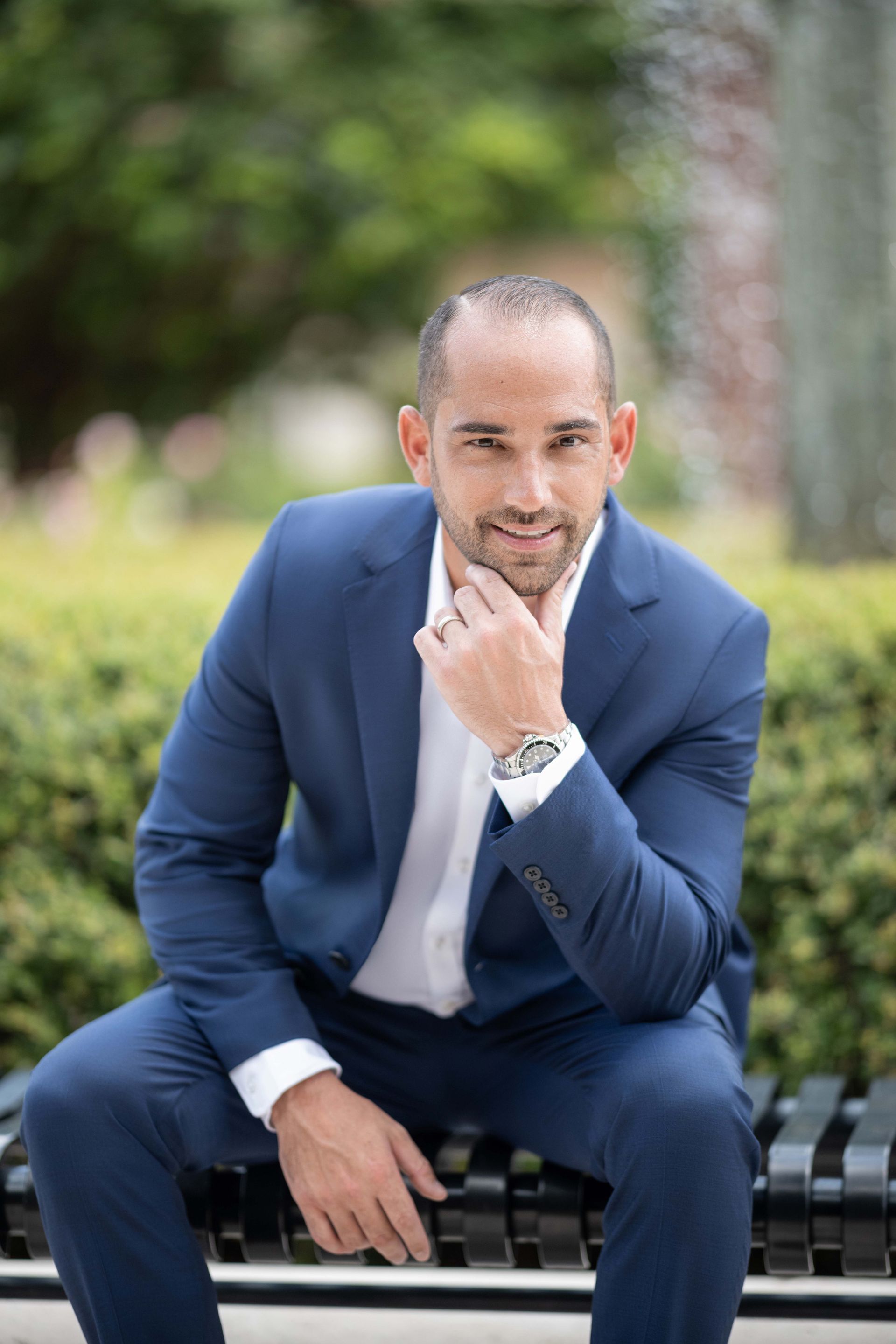 Man in a blue suit sitting on a bench outside, hand on his chin, smiling.