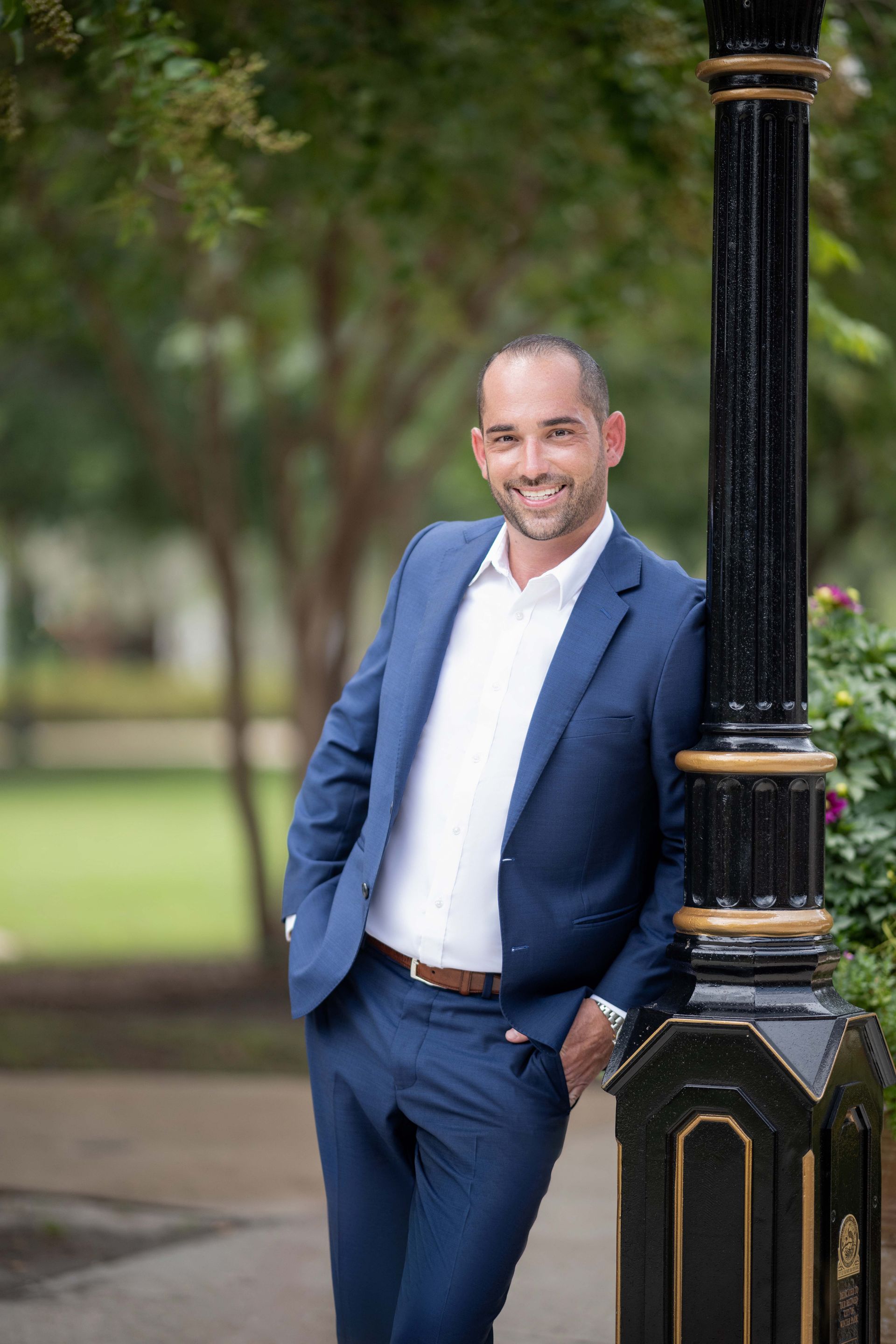 Man in a blue suit smiles, leaning against a black lamppost in an outdoor park setting.