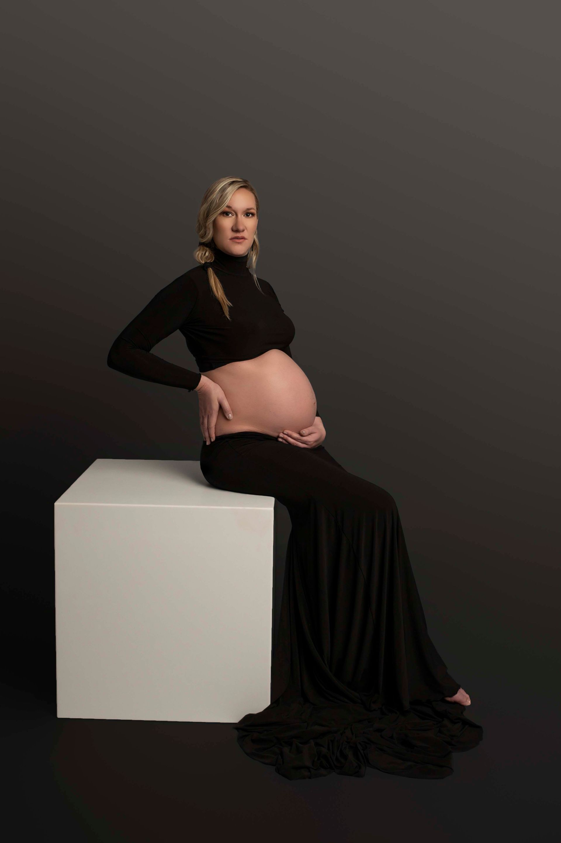 Pregnant woman in black dress sits on a white cube, holding belly, against dark gray backdrop.