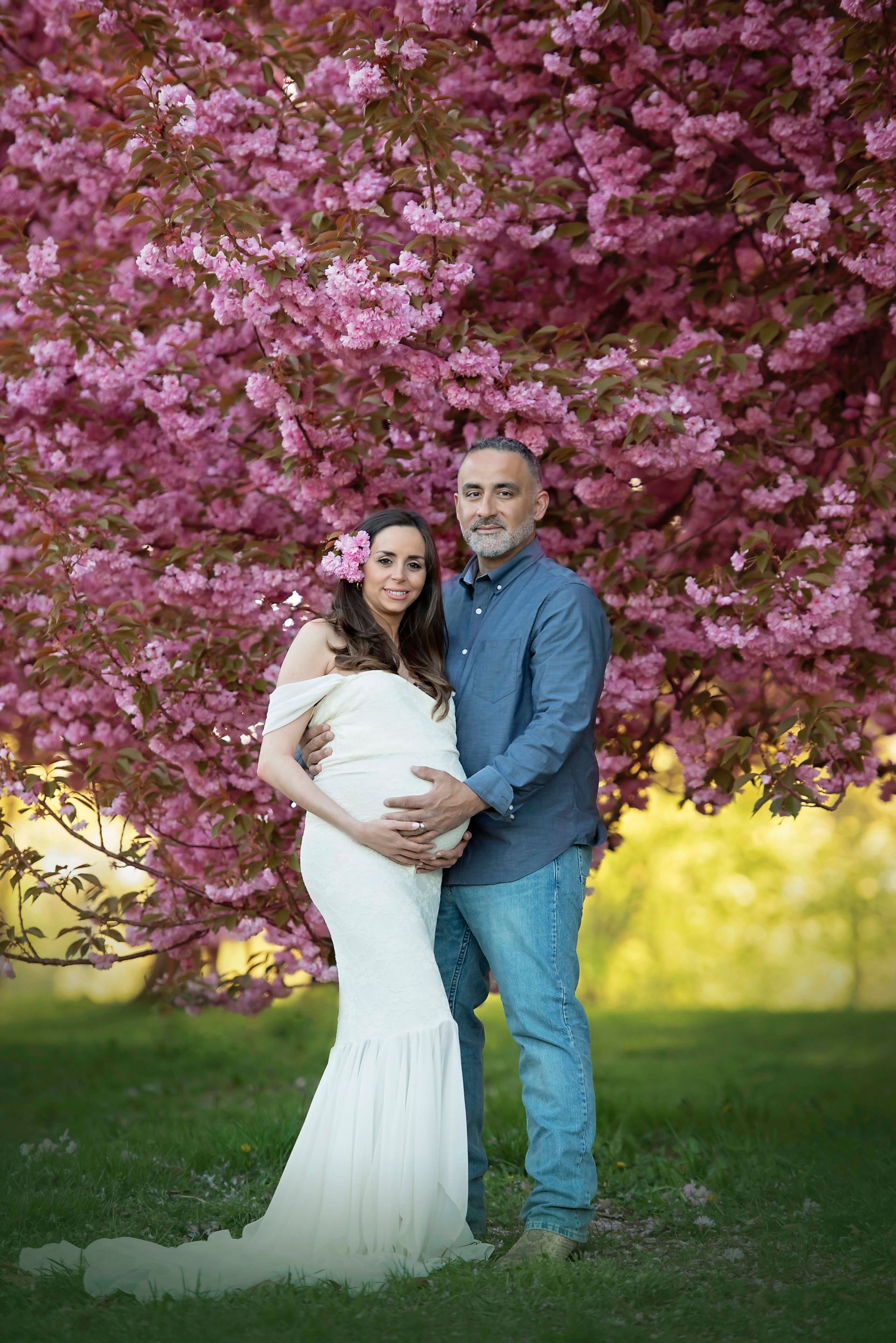 Pregnant woman and man pose in front of a blooming pink tree. Woman holds belly, wearing white gown, man wears blue shirt.