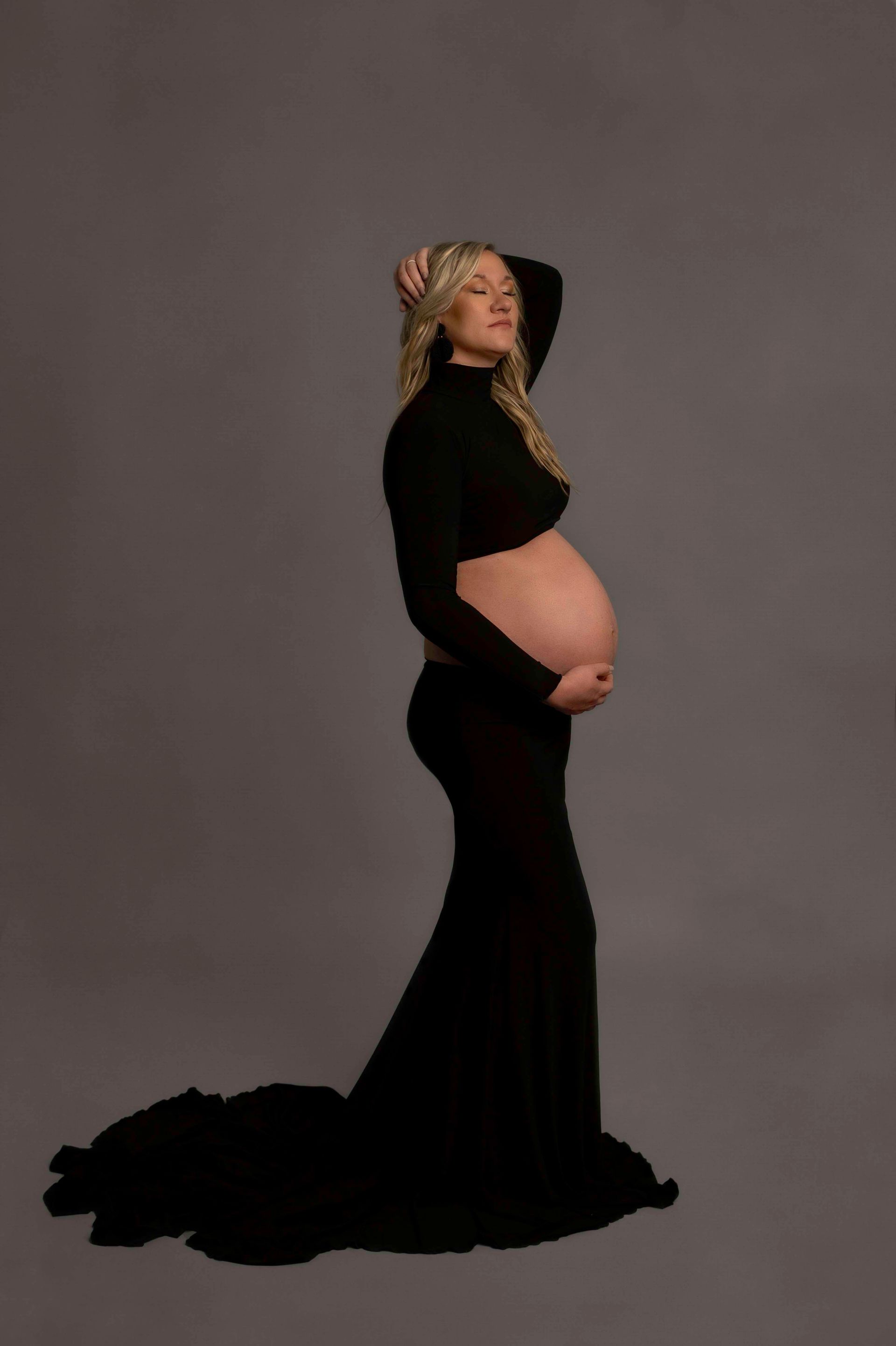 Pregnant woman in black dress, hands on belly, head tilted back, against gray backdrop.