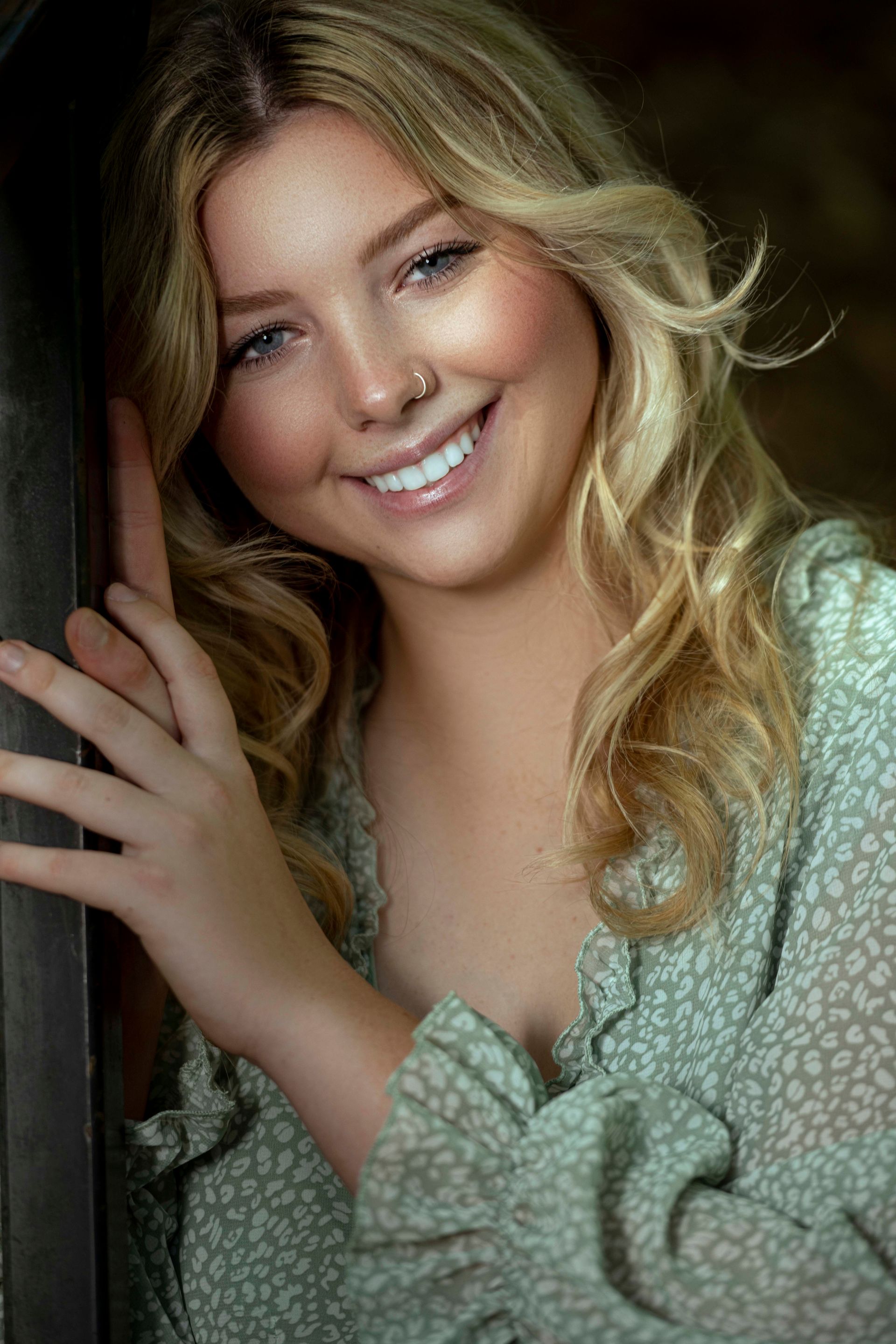 Blonde woman smiling, leaning against a wooden structure. She wears a green floral top.