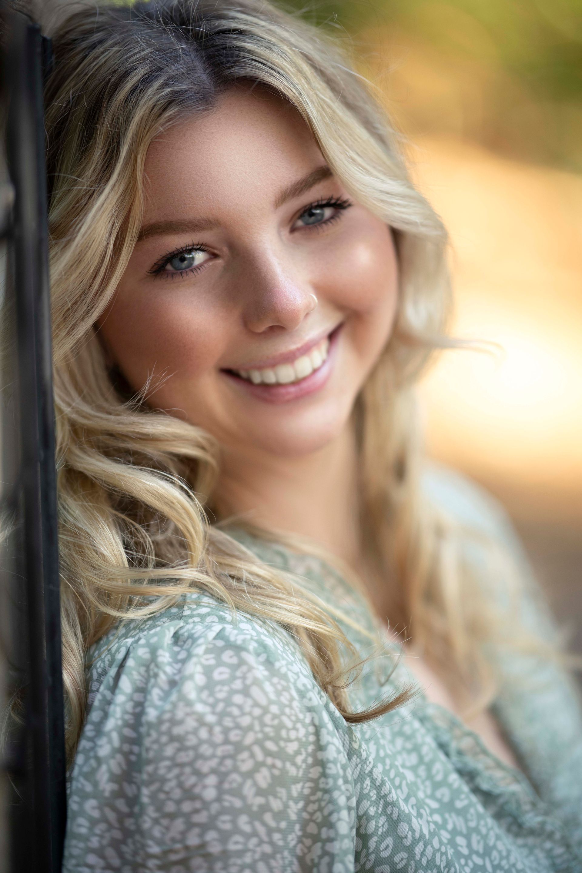 Blonde woman smiles, leaning on a dark post, wearing a green dress outdoors.