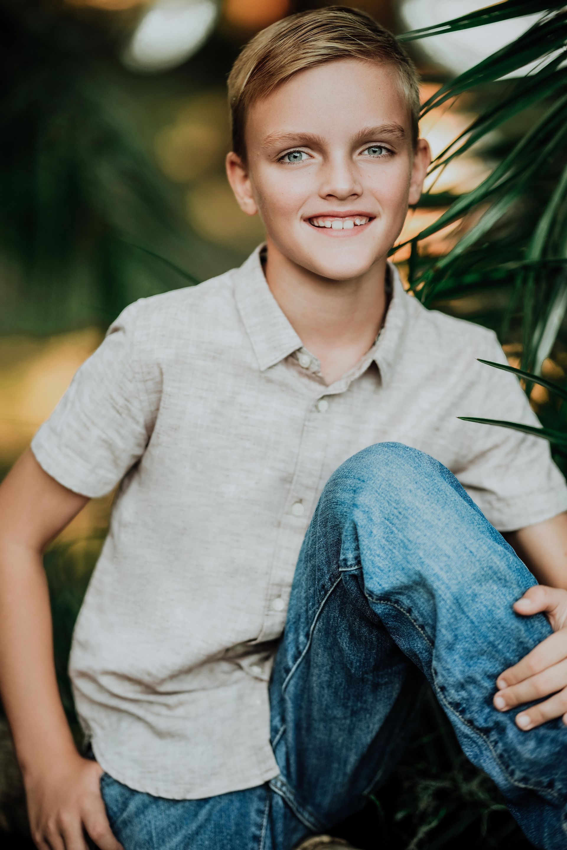Smiling person in a light shirt and jeans seated outdoors beside green plants.