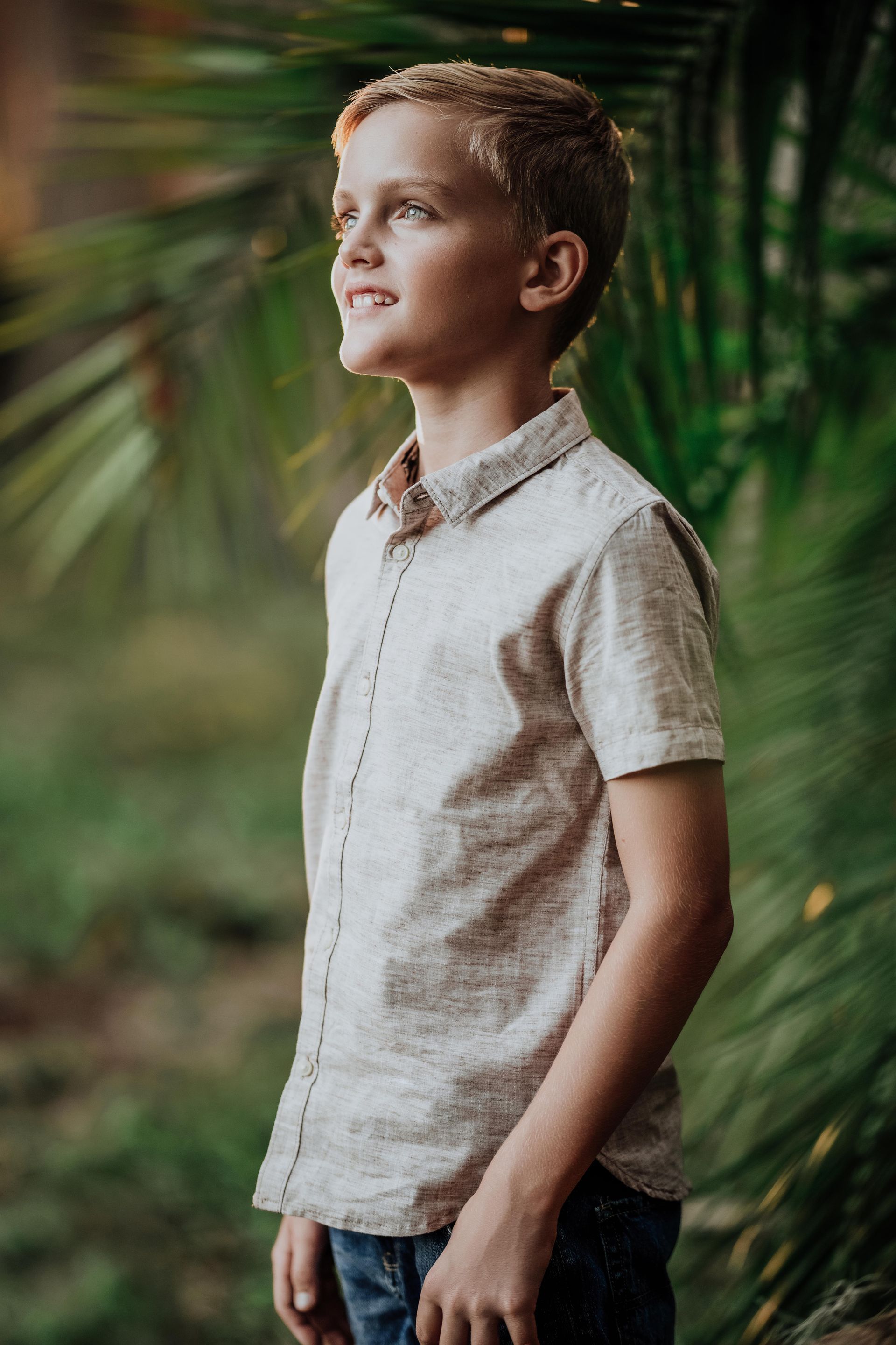Boy standing outdoors among green leaves, wearing a light shirt and looking to the side.