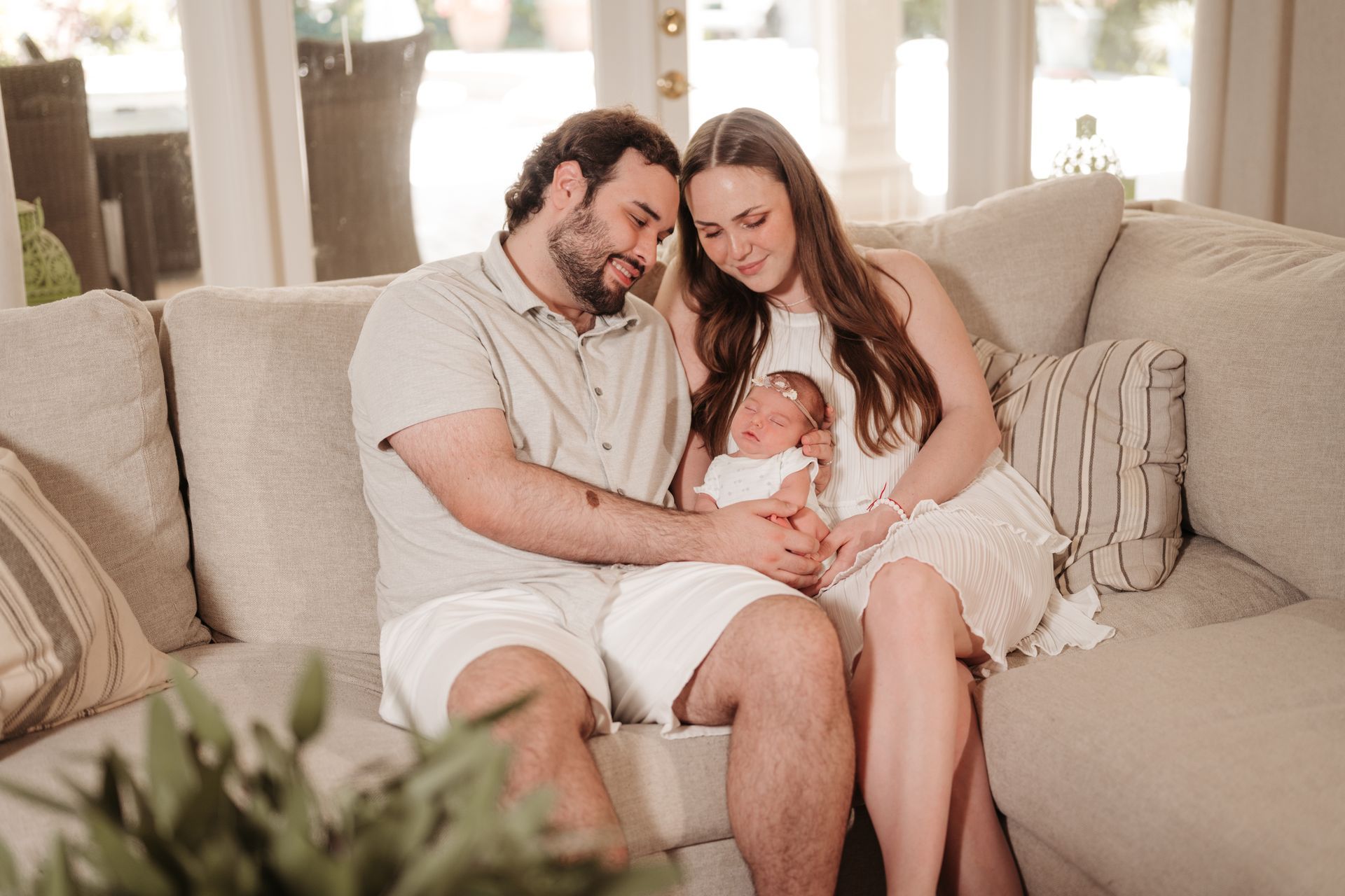 Couple sitting on a beige sofa, smiling and holding a baby in a bright living room