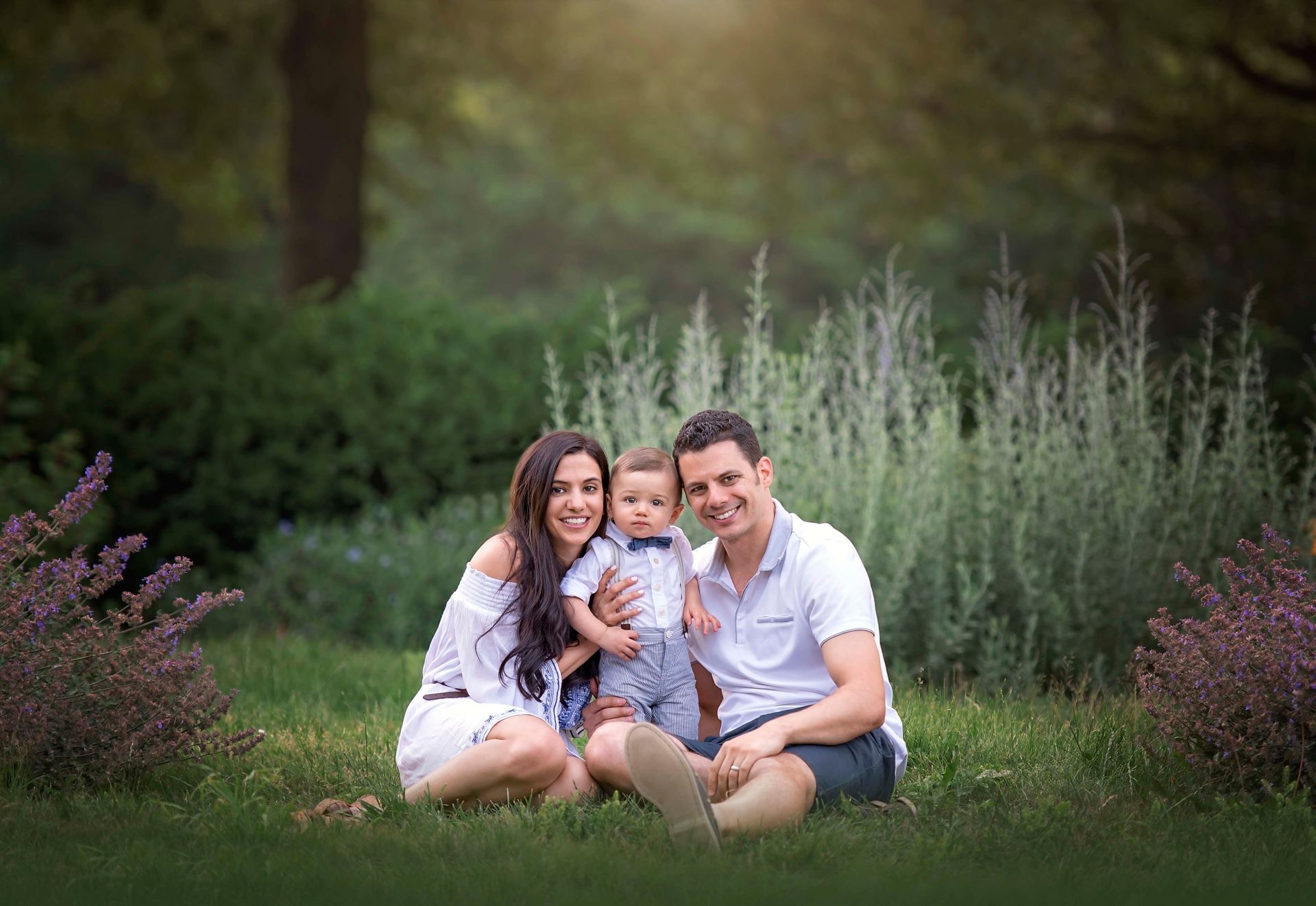 Three people sitting on grass in a garden, smiling and holding a small dog.
