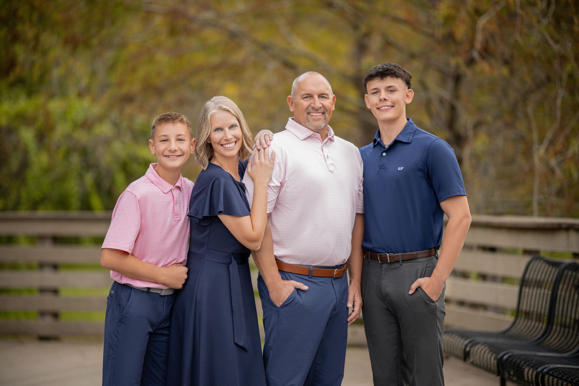 Four people posing together outdoors by a wooden fence and bench, smiling in casual clothes.