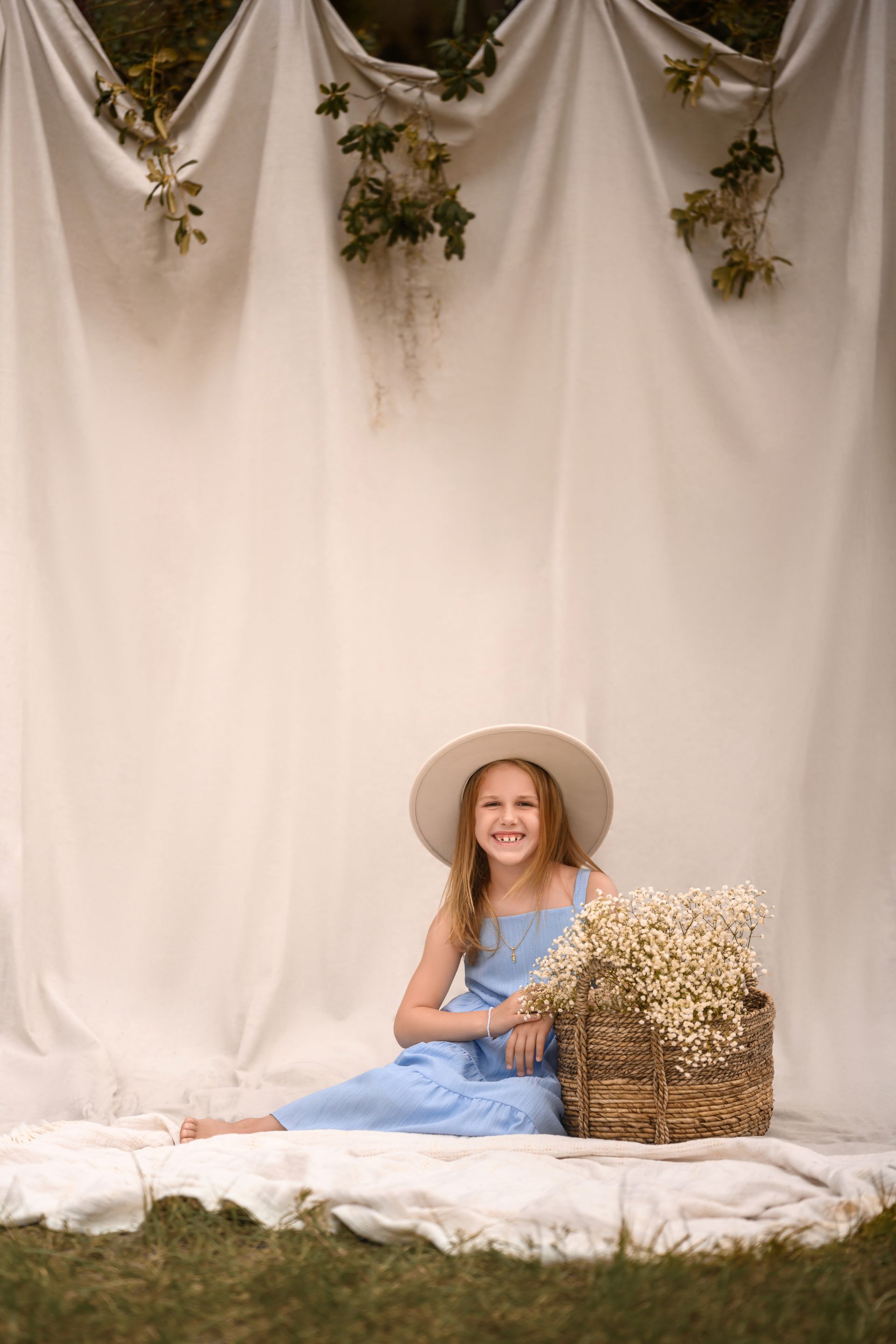 Woman in a blue dress and straw hat sitting on a white blanket beside a woven basket under draped fabric and greenery.
