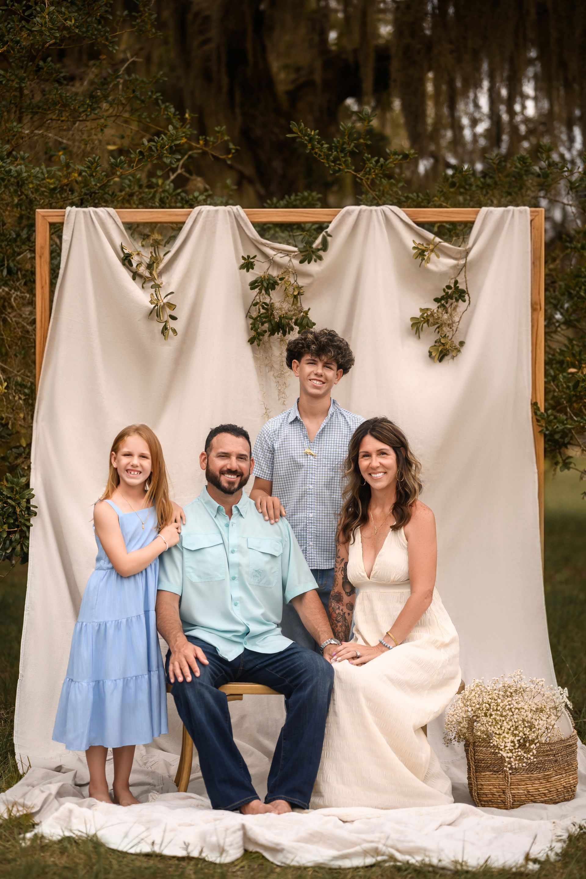 Family portrait in front of a draped backdrop outdoors, with four people seated and standing, smiling.