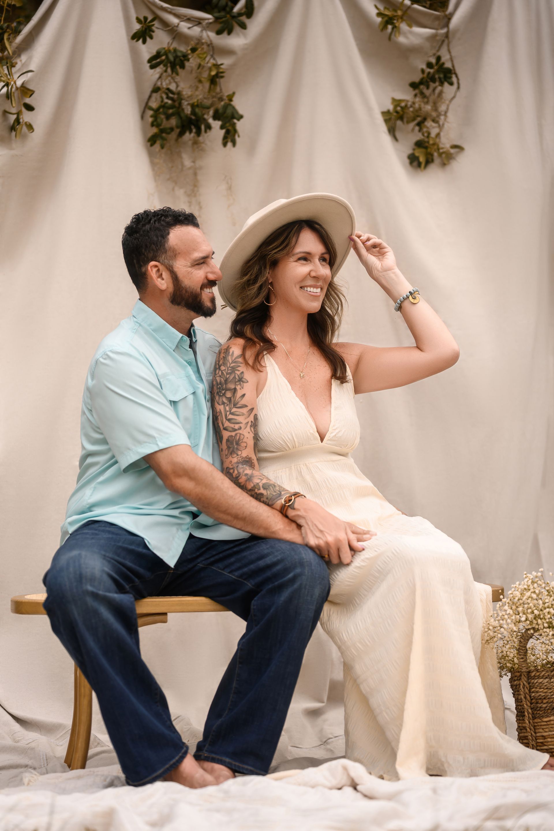 Couple seated on a bench at a rustic wedding setting, woman in white dress and hat, man in blue shirt