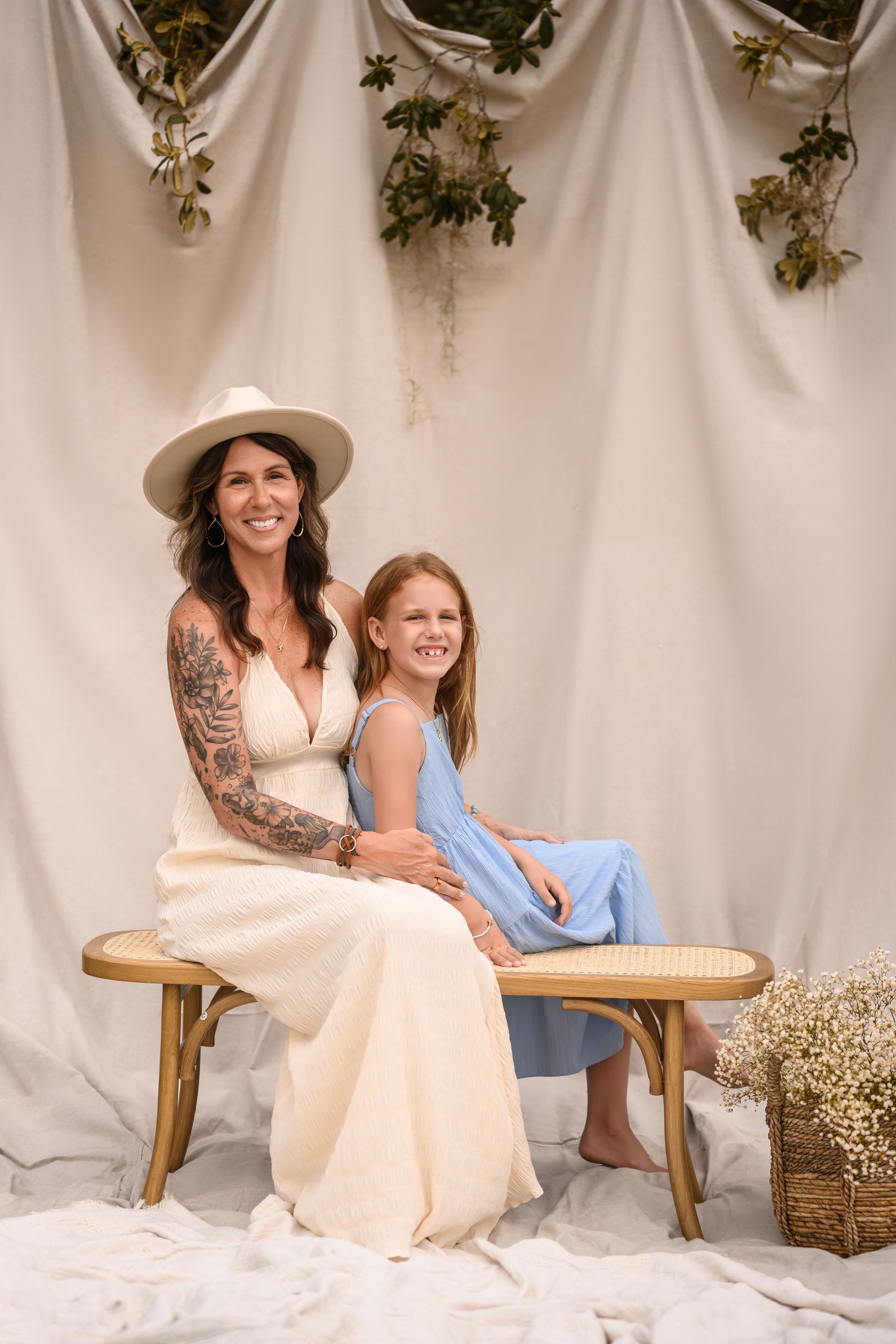 Woman and girl sitting on a bench in a cream-draped floral setting, both smiling in light dresses