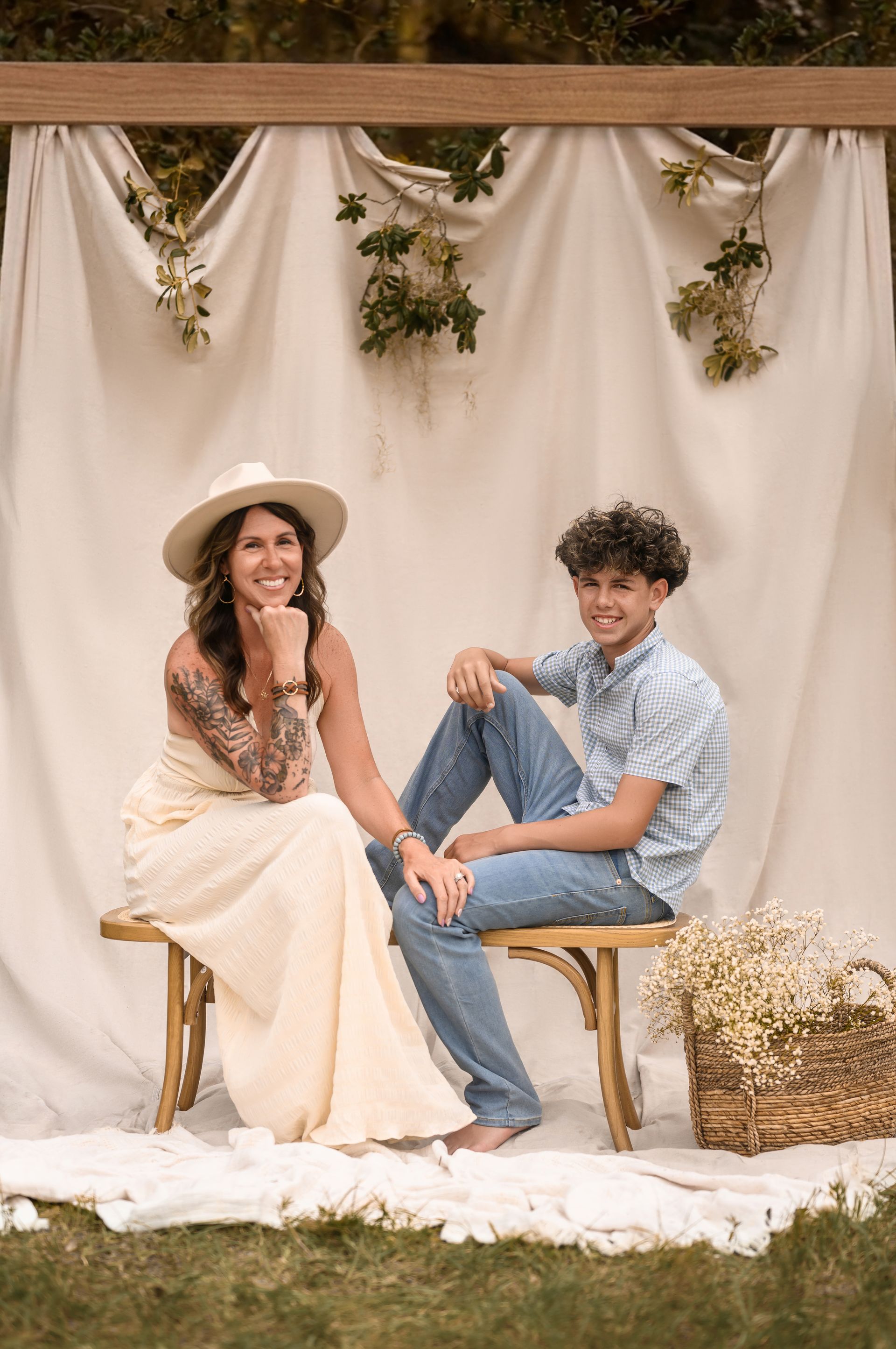 Two people seated on wooden chairs before a cream backdrop with greenery and white fabric draping