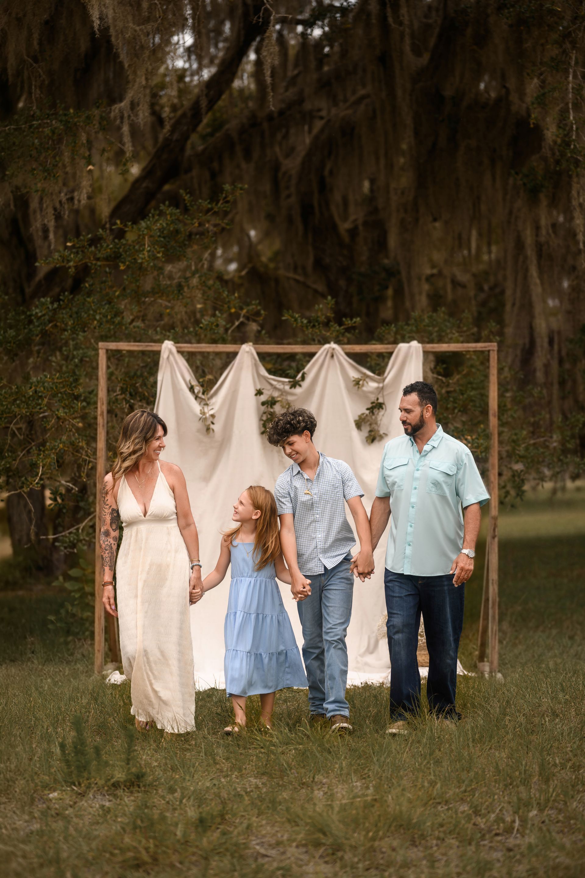 Family of four standing outdoors under a rustic frame with white draped fabric, holding hands in a grassy field.