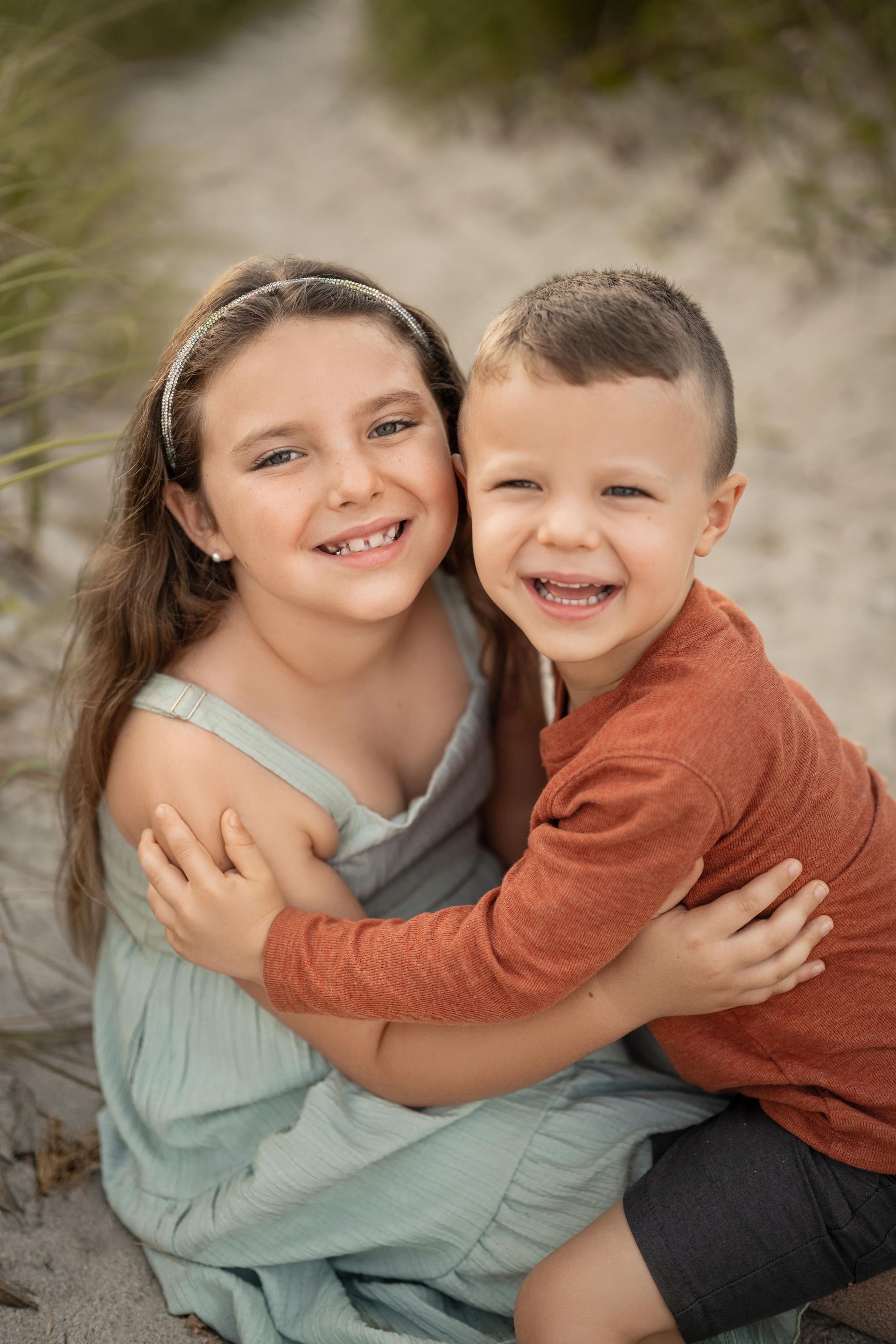Two smiling children hugging outdoors, one in a green dress and one in an orange sweater.