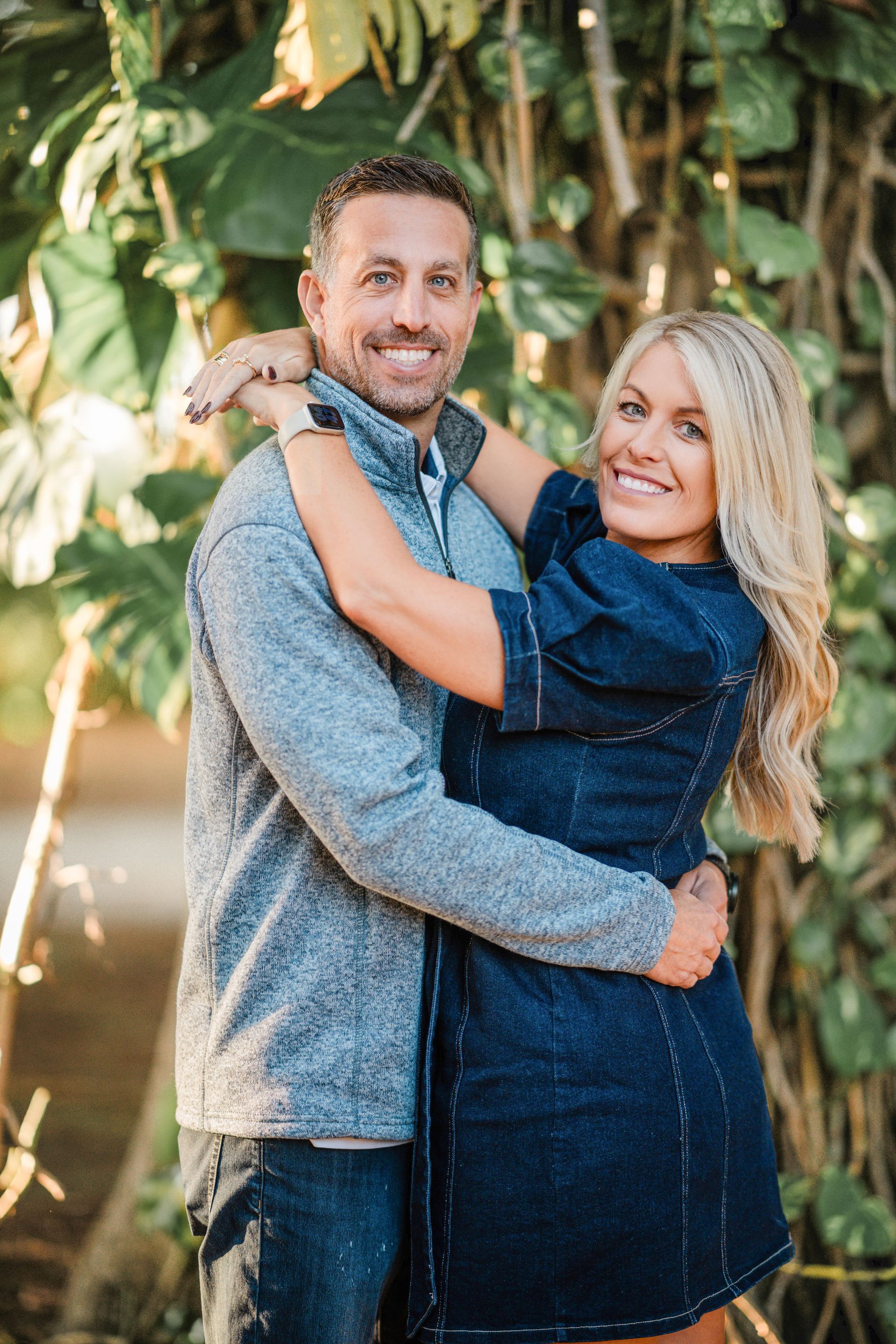 Smiling couple hugging outdoors in front of lush greenery, both in blue outfits.