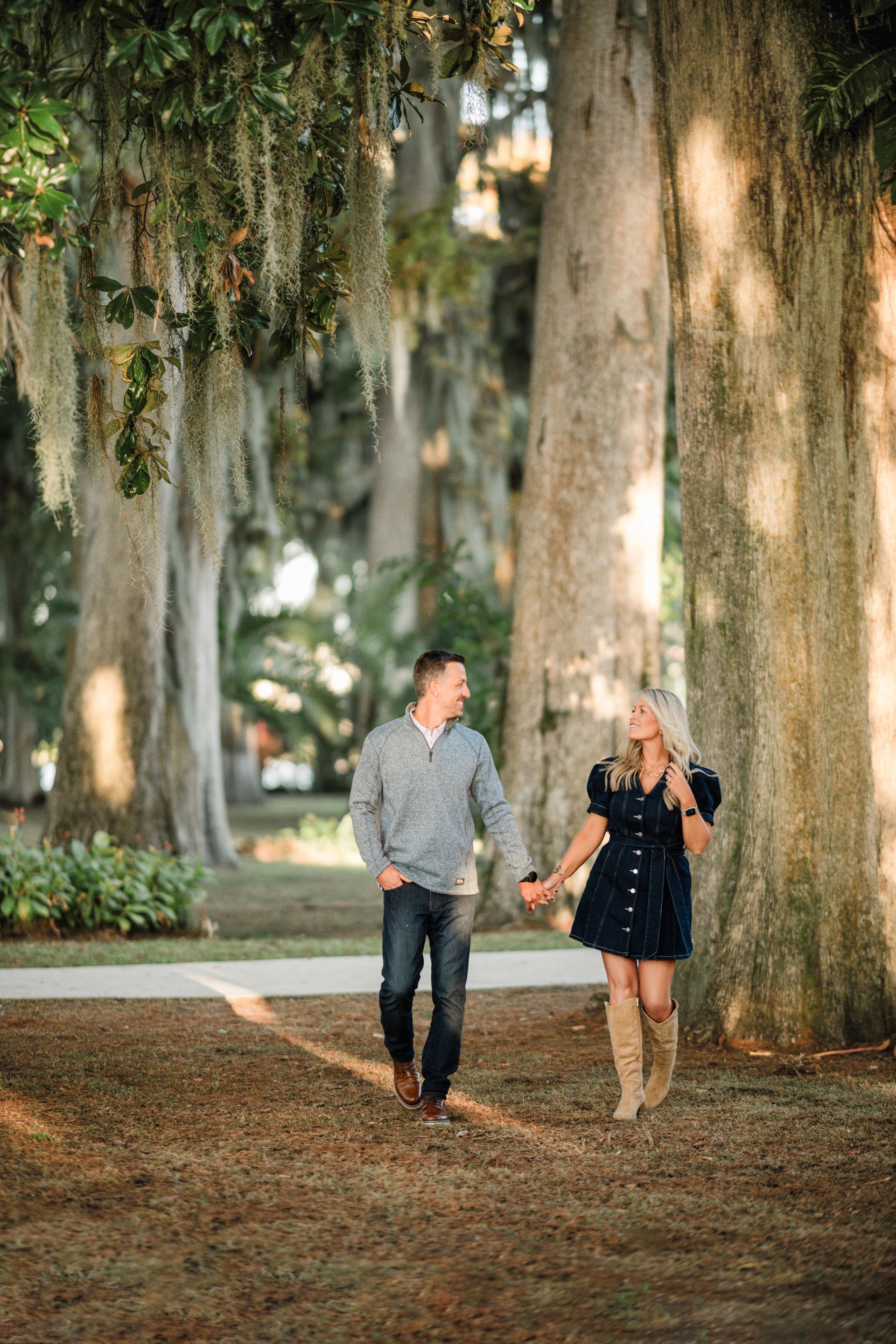 Two people walking hand in hand on a tree-lined path in a park, with large oaks in the background