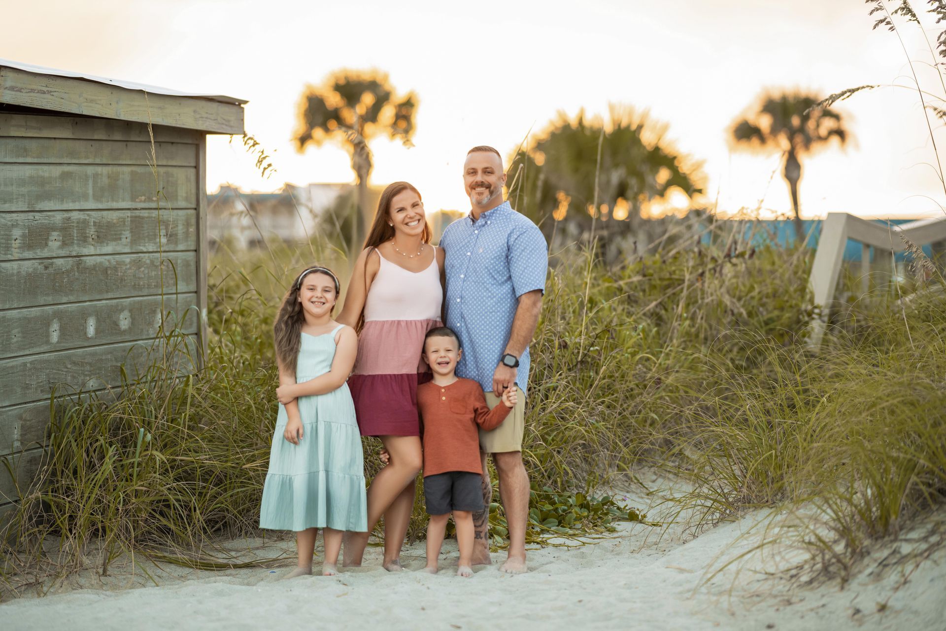 Family of four standing on a sandy beach at sunset near dunes and a small hut.