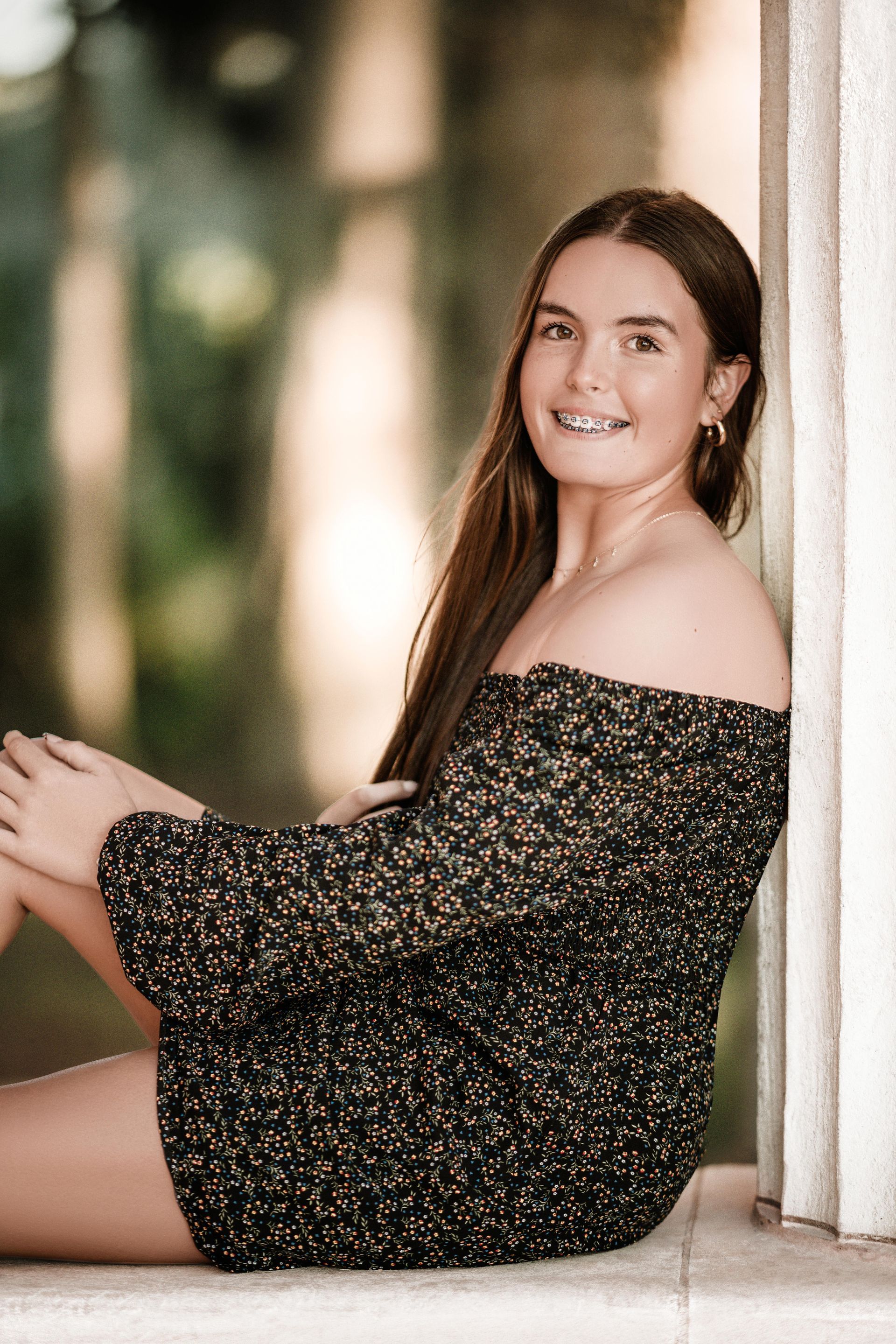 Young person in a black off-shoulder dress sitting by a white column, smiling outdoors